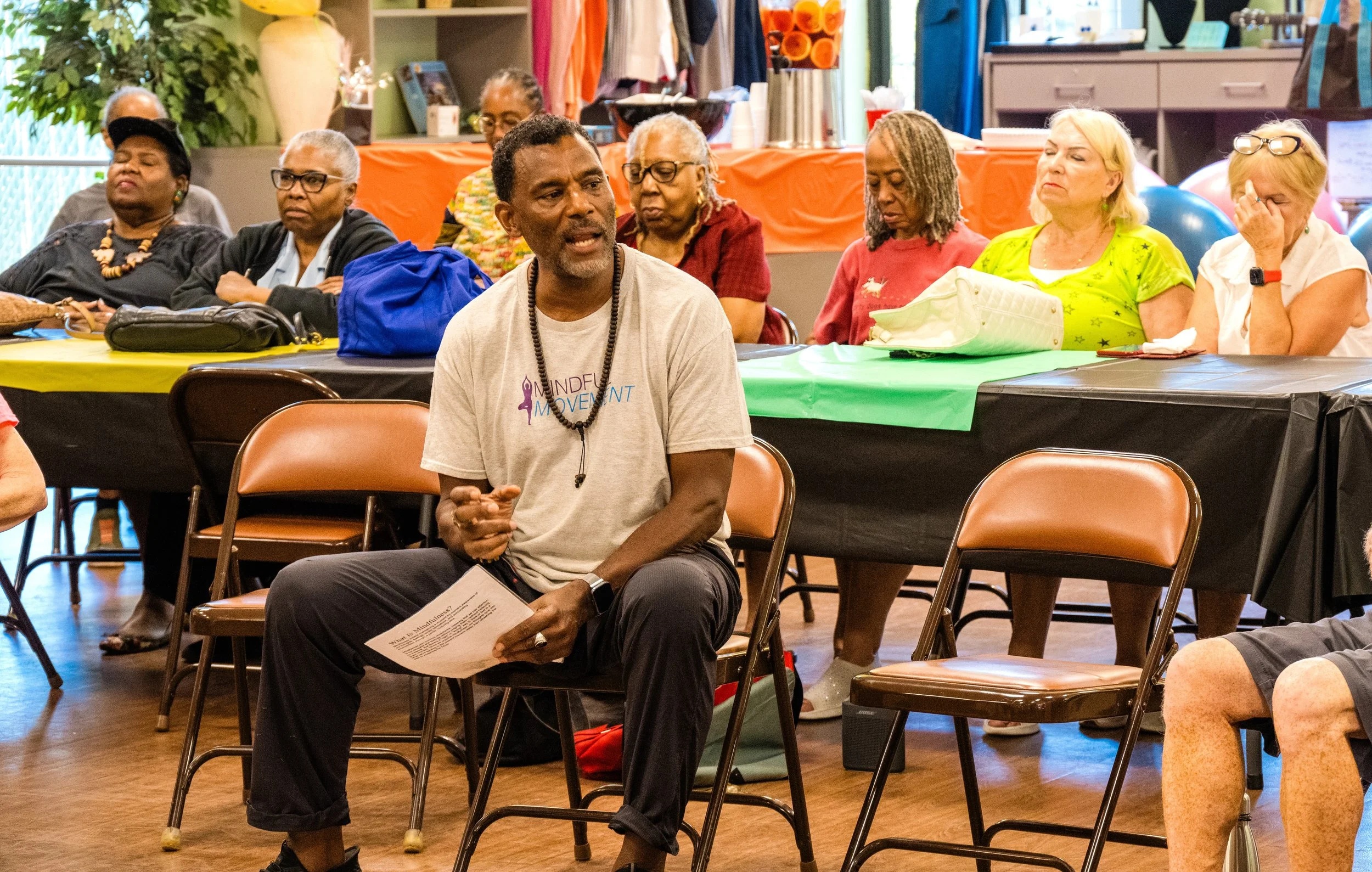 A man in a gray t-shirt and black pants sitting on a chair, speaking to a group of older women, some of whom appear to be taking notes or listening attentively, in a colorful room with tables covered in green, yellow, orange, and black tablecloths, and balloons in the background.