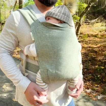 A person carrying a baby in a soft, green baby carrier outdoors in a wooded area during daytime.