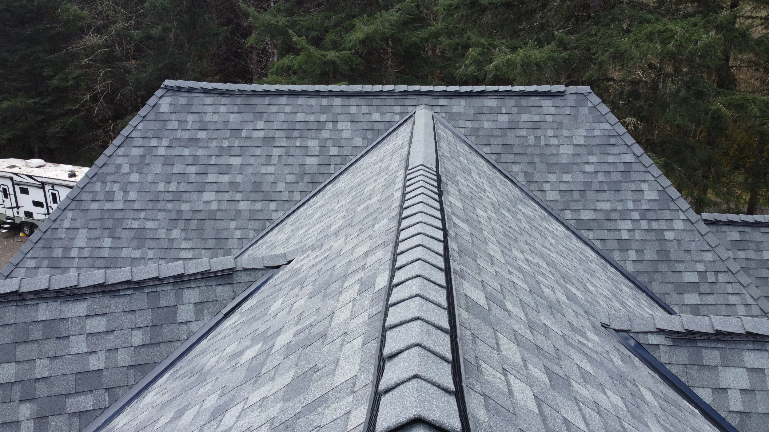 Aerial view of a gray shingled roof with a ridge cap, surrounded by trees and a section of a white RV on the left side.