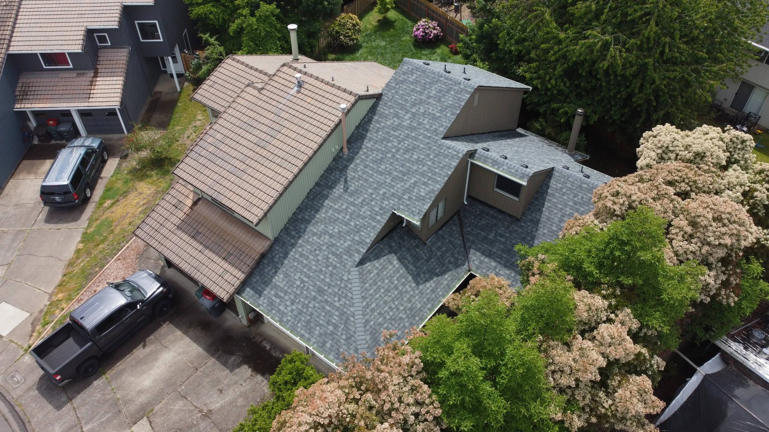 An aerial view of a residential house with a dark gray roof, a small attached shed or extension, surrounded by trucks and cars, with green trees and flowering bushes in a suburban neighborhood.