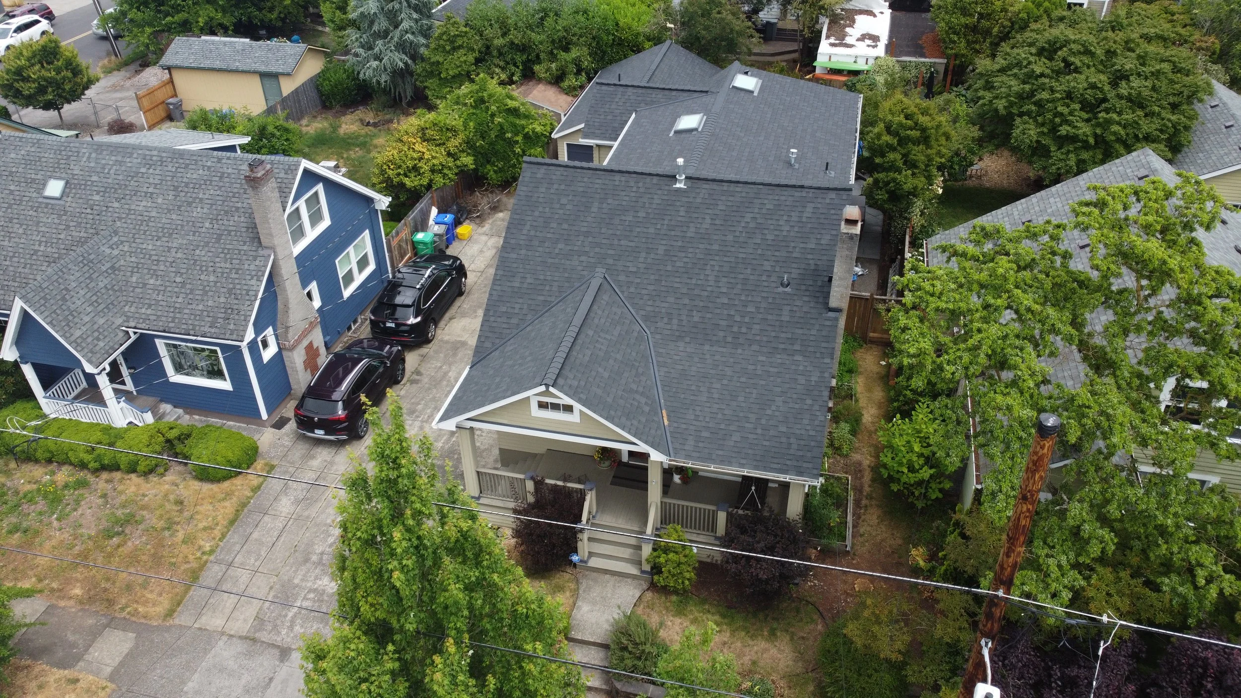 An aerial view of a neighborhood showing houses, cars parked in driveways, trees, and utility poles with wires.