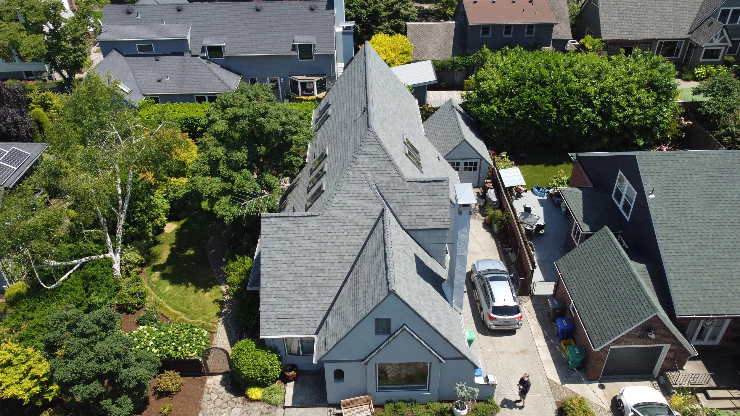 Aerial view of a suburban neighborhood with multiple houses, trees, and a backyard. Prominent house has a light grey roof, driveway, and a person walking near the sidewalk.