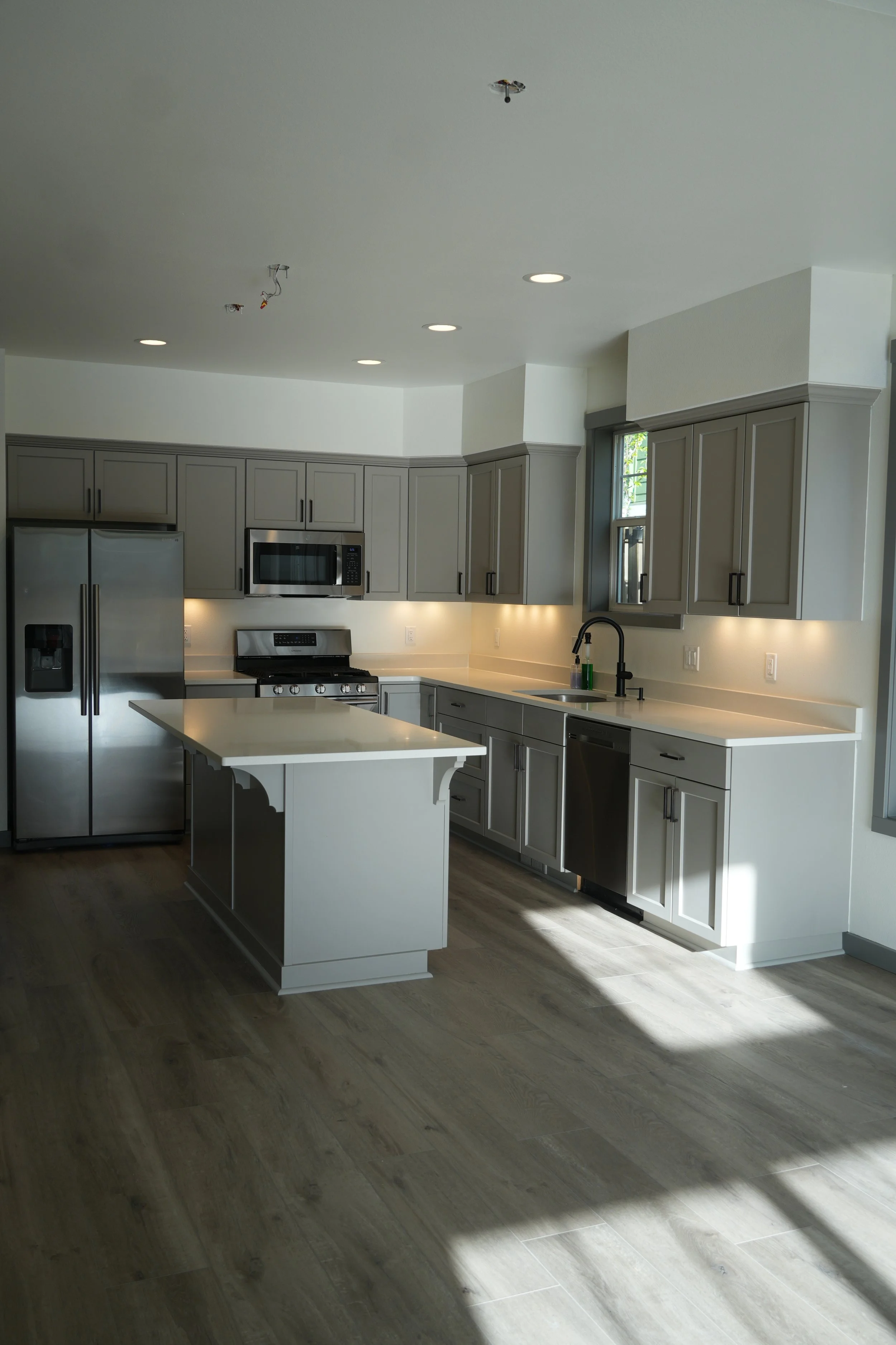 Modern kitchen with gray cabinets, stainless steel appliances, including refrigerator, microwave, oven, and dishwasher, with light-colored countertops and wood flooring.