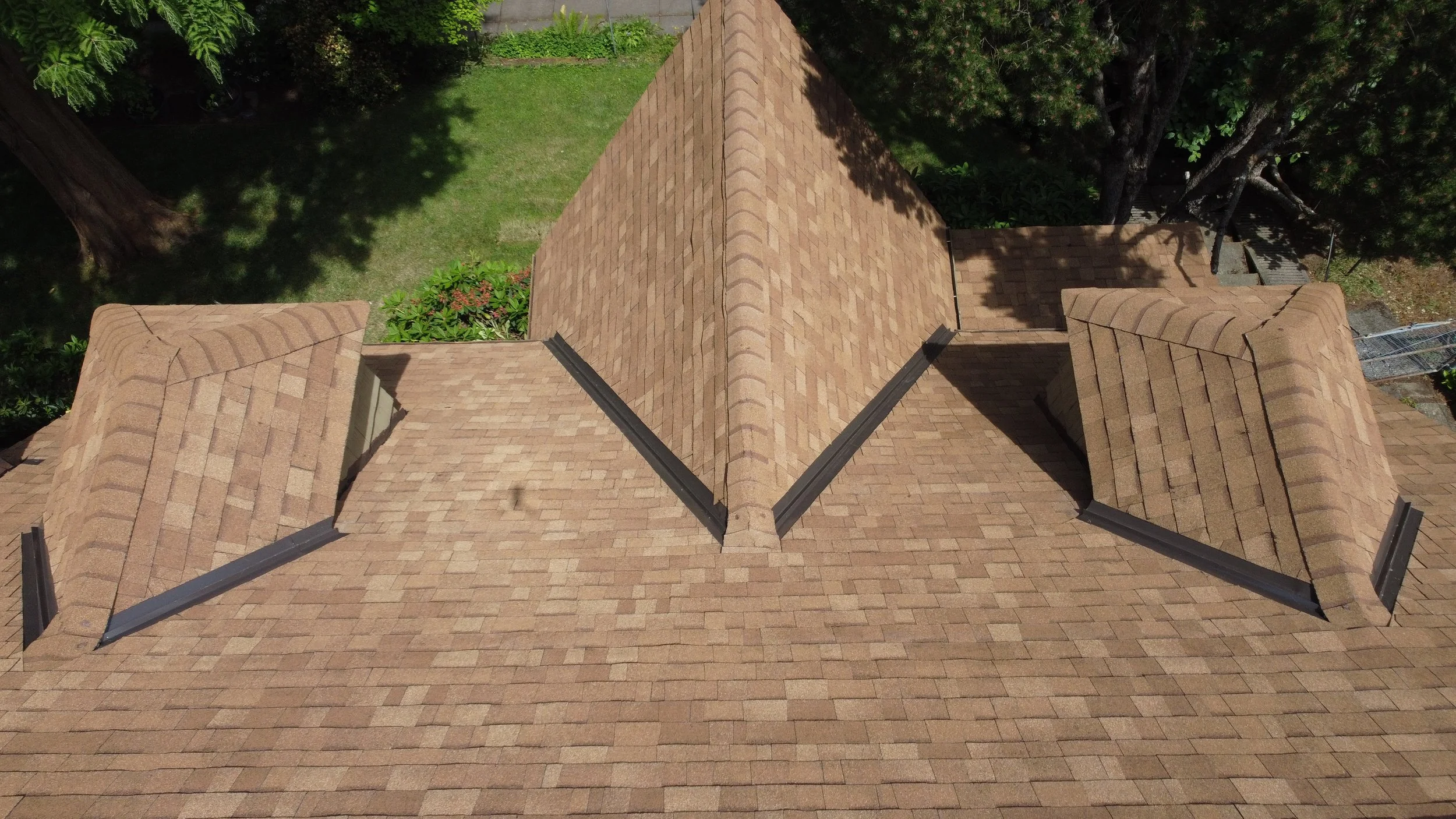 An aerial view of a house roof with multiple gabled sections covered in tan shingles, with trees and greenery surrounding the house.