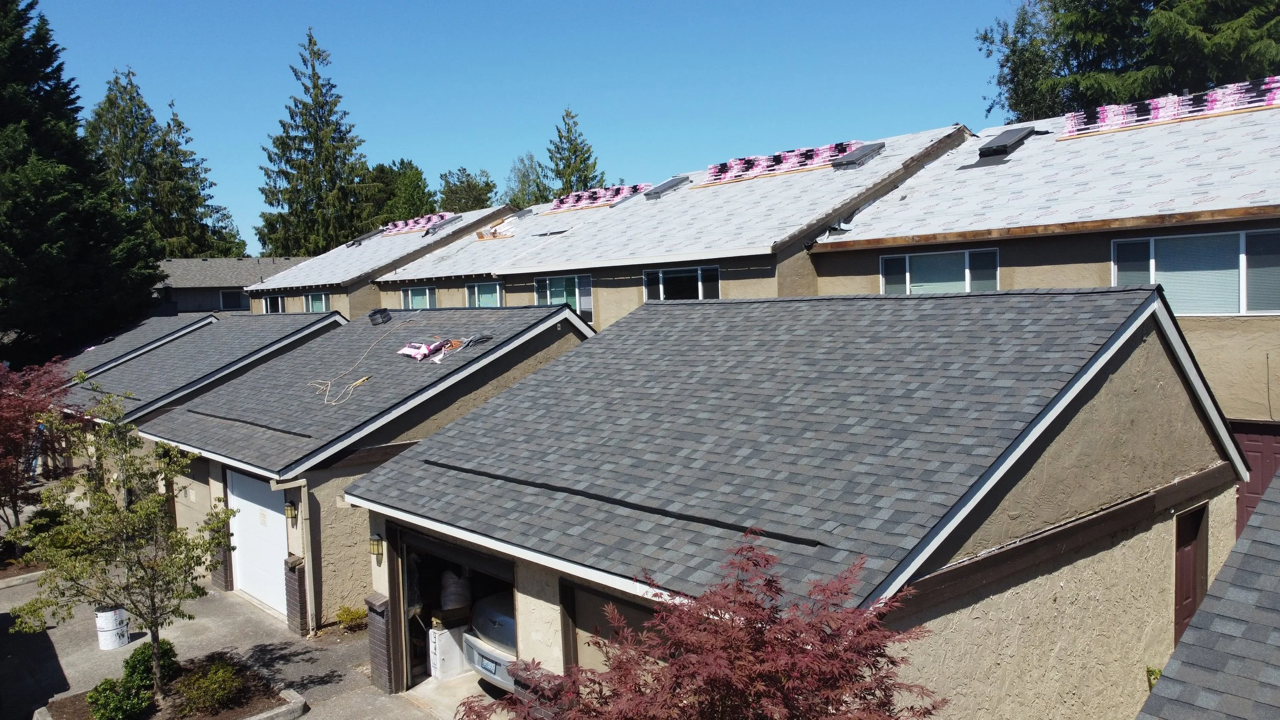 Row of houses with roofs under construction, some with new shingles and others still needing roofing work, surrounded by trees on a sunny day.