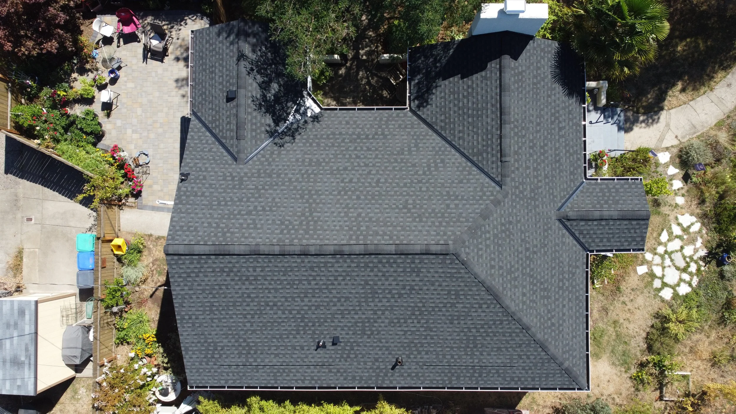 An aerial view of a house with a dark gray shingled roof, surrounded by a backyard with a patio, garden, and various outdoor furniture and plants.