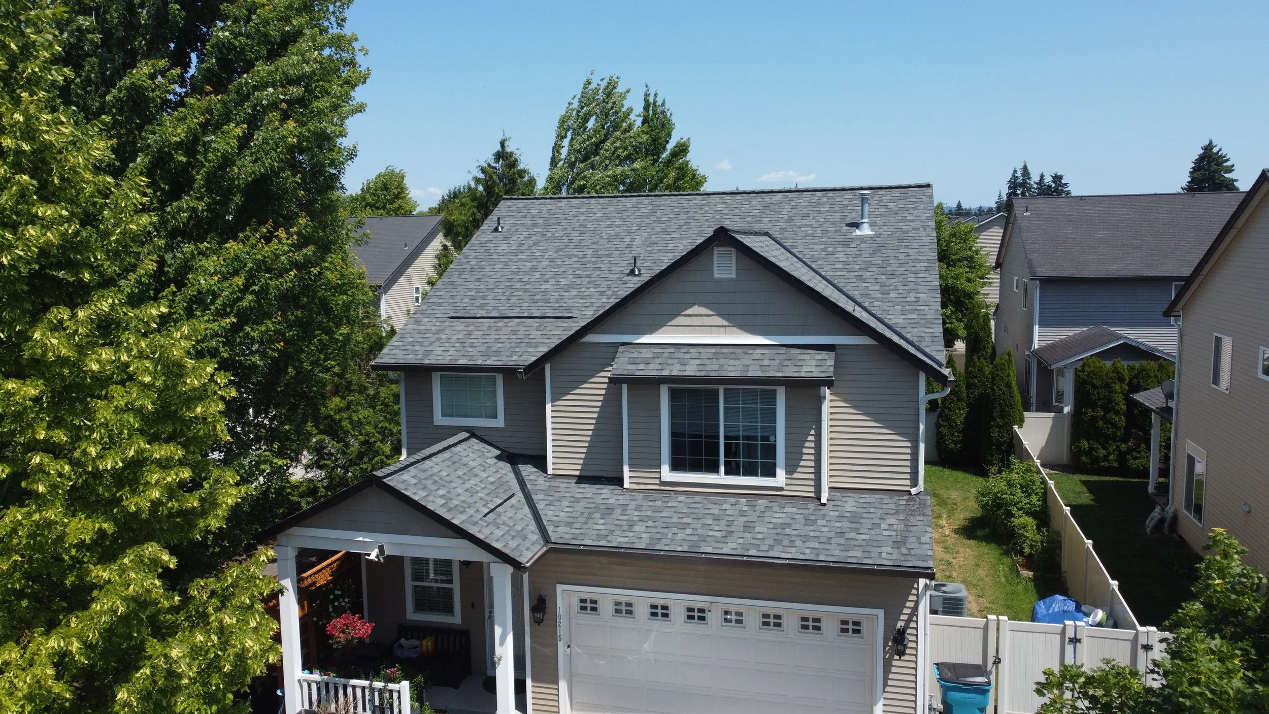 A two-story suburban house with beige siding and gray roof, surrounded by trees and neighboring houses, under a blue sky.