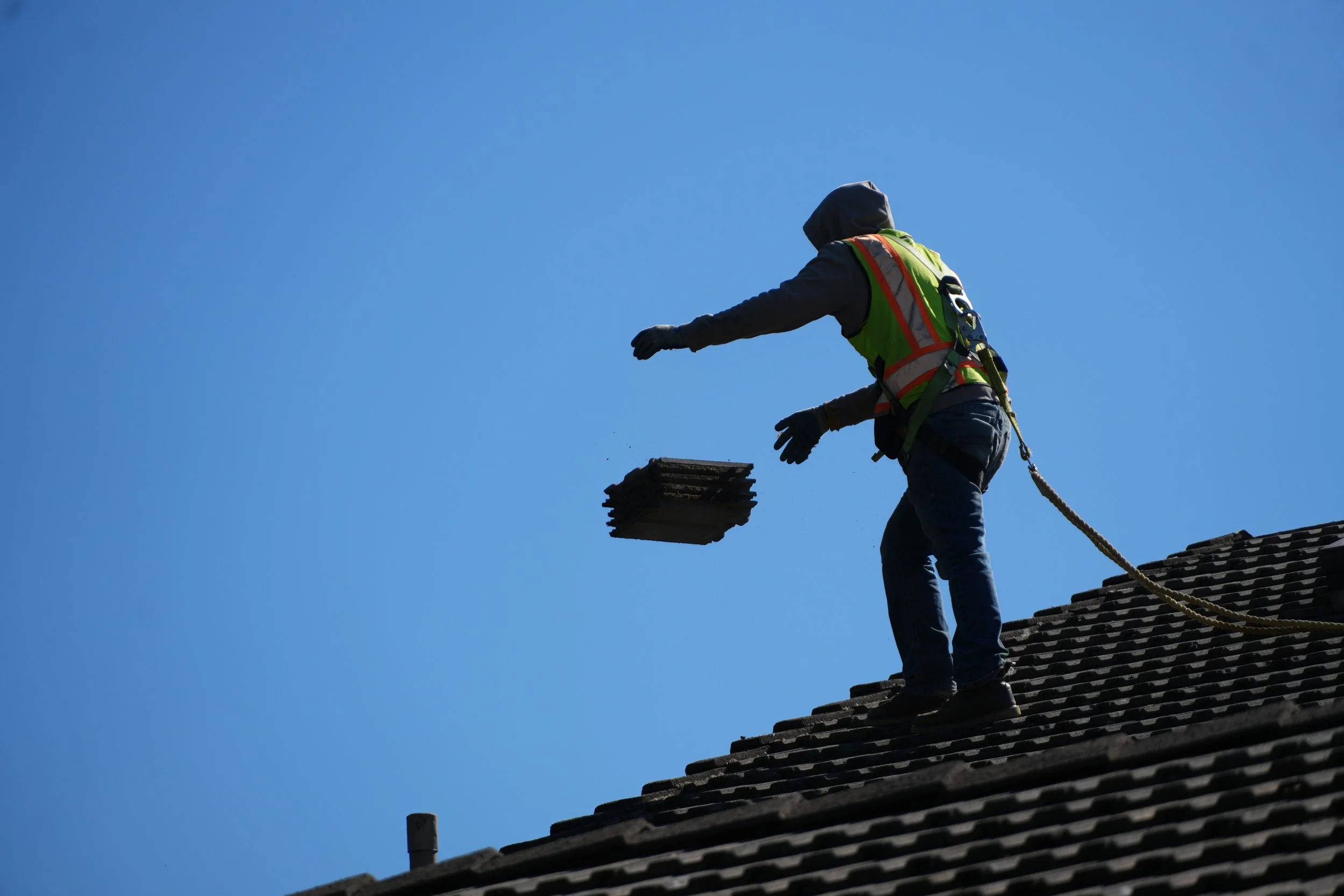 Worker on roof, wearing safety gear, tossing tiles in the air against a clear blue sky.