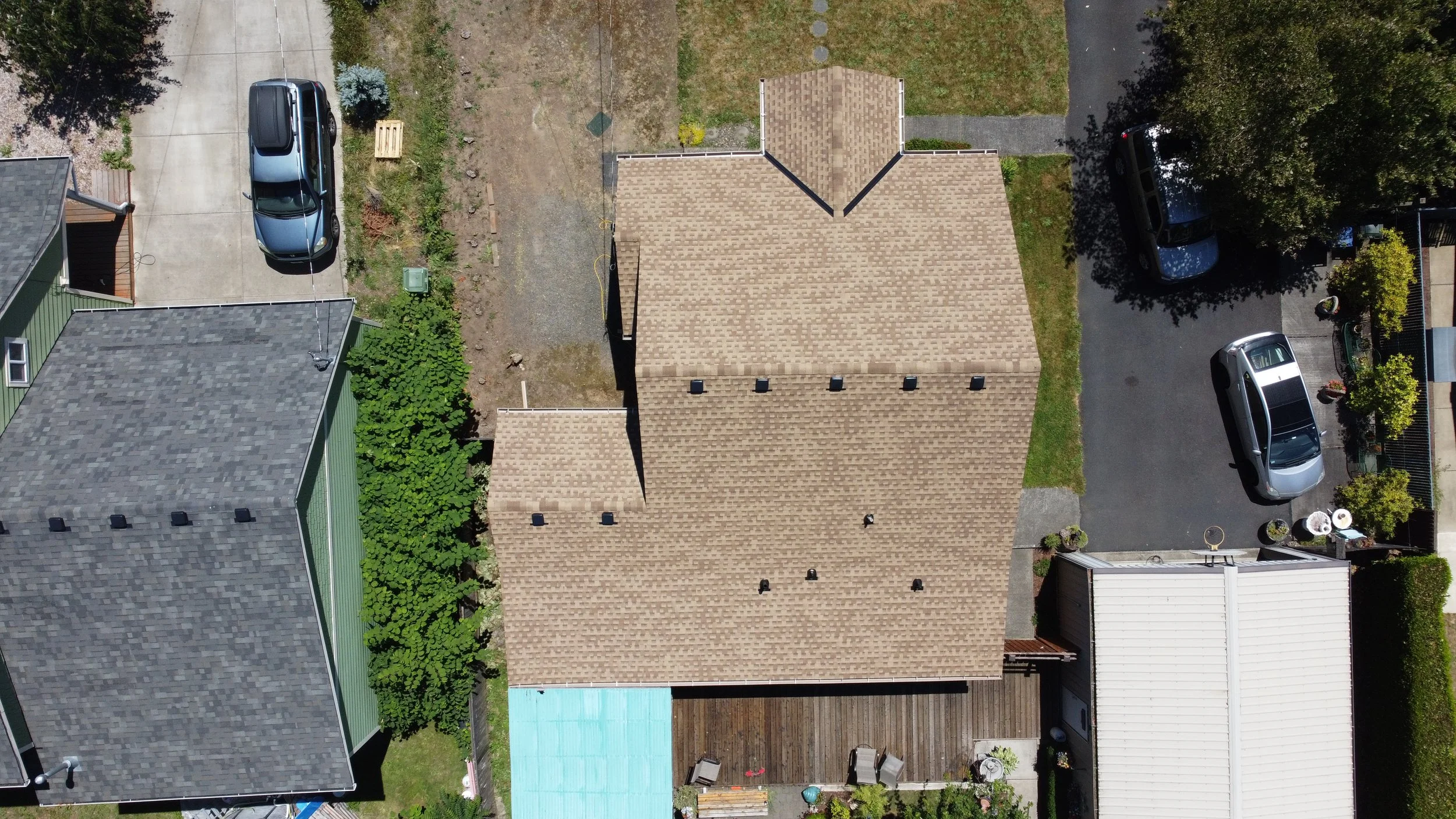 An aerial view of a house with a brown shingled roof, surrounded by a driveway with parked cars, a backyard with patio furniture, and neighboring houses with various roof styles.