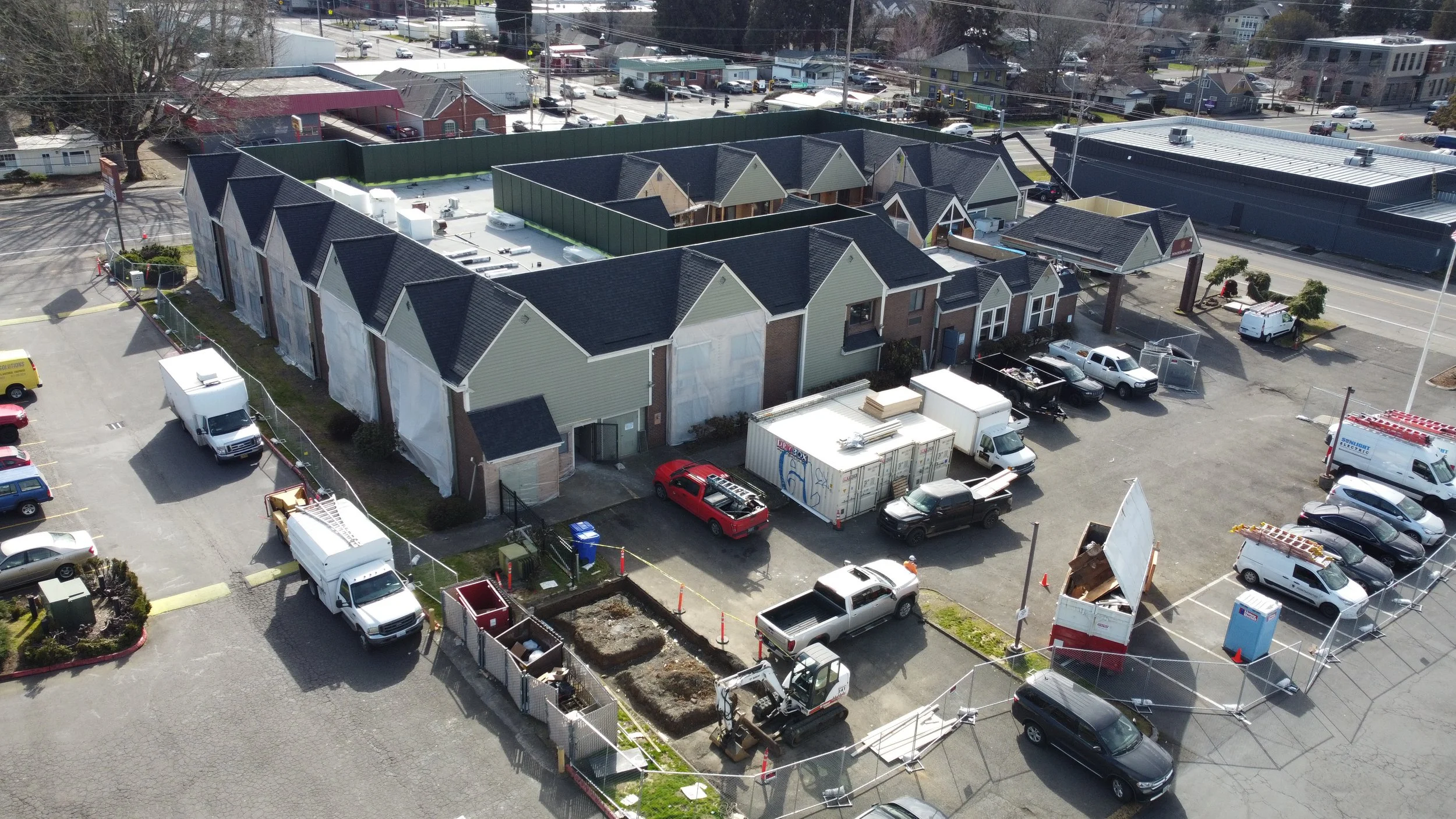 Construction site with new townhomes under construction, surrounded by parked cars, construction equipment, and a small fenced area, viewed from above.