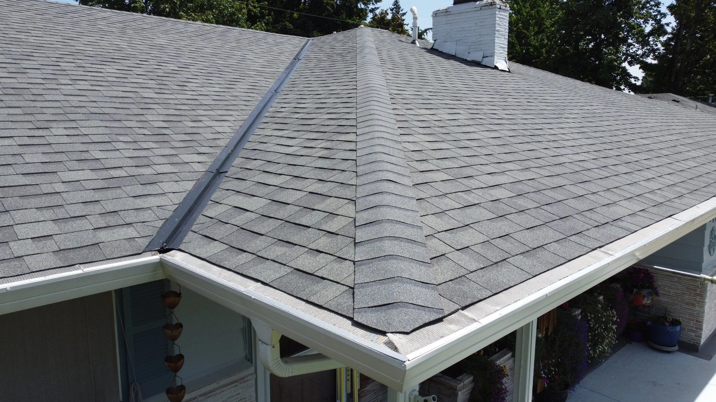 A house roof with dark gray asphalt shingles, featuring a central ridge cap, a white chimney, and a metal ridge vent, surrounded by green trees.