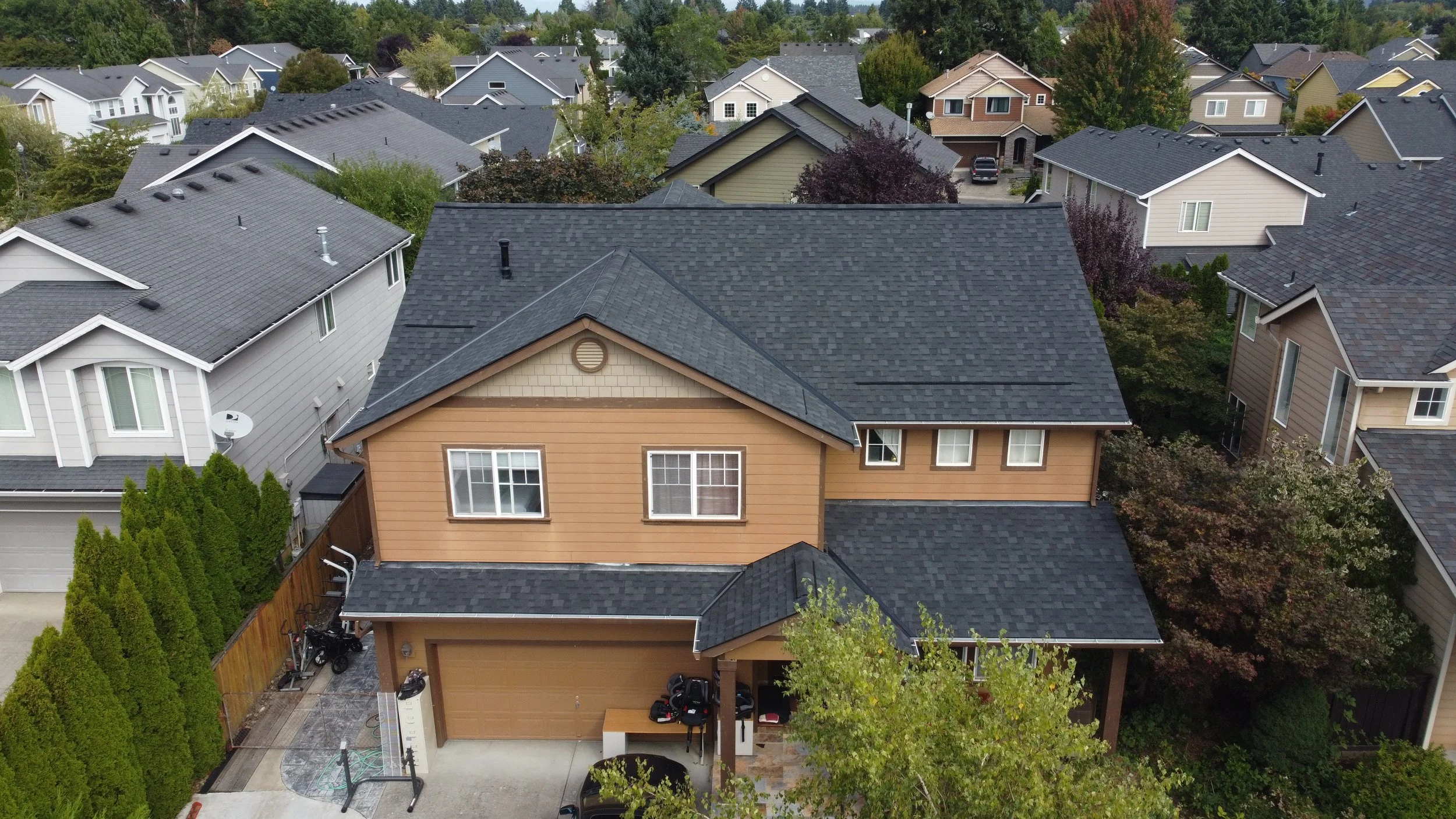 An aerial view of a two-story house with beige siding, black roof, and attached garage surrounded by other similar houses in a suburban neighborhood with trees.