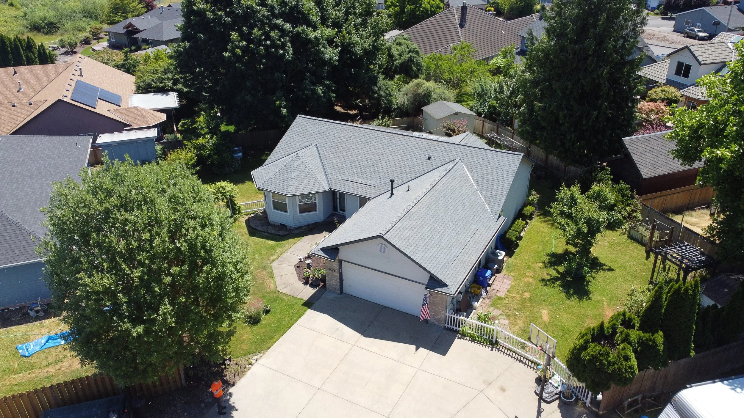 An aerial view of a suburban house with a gray roof, surrounded by green trees and a backyard. The house has a driveway with a basketball hoop and a small fenced yard with various plants and garden features.