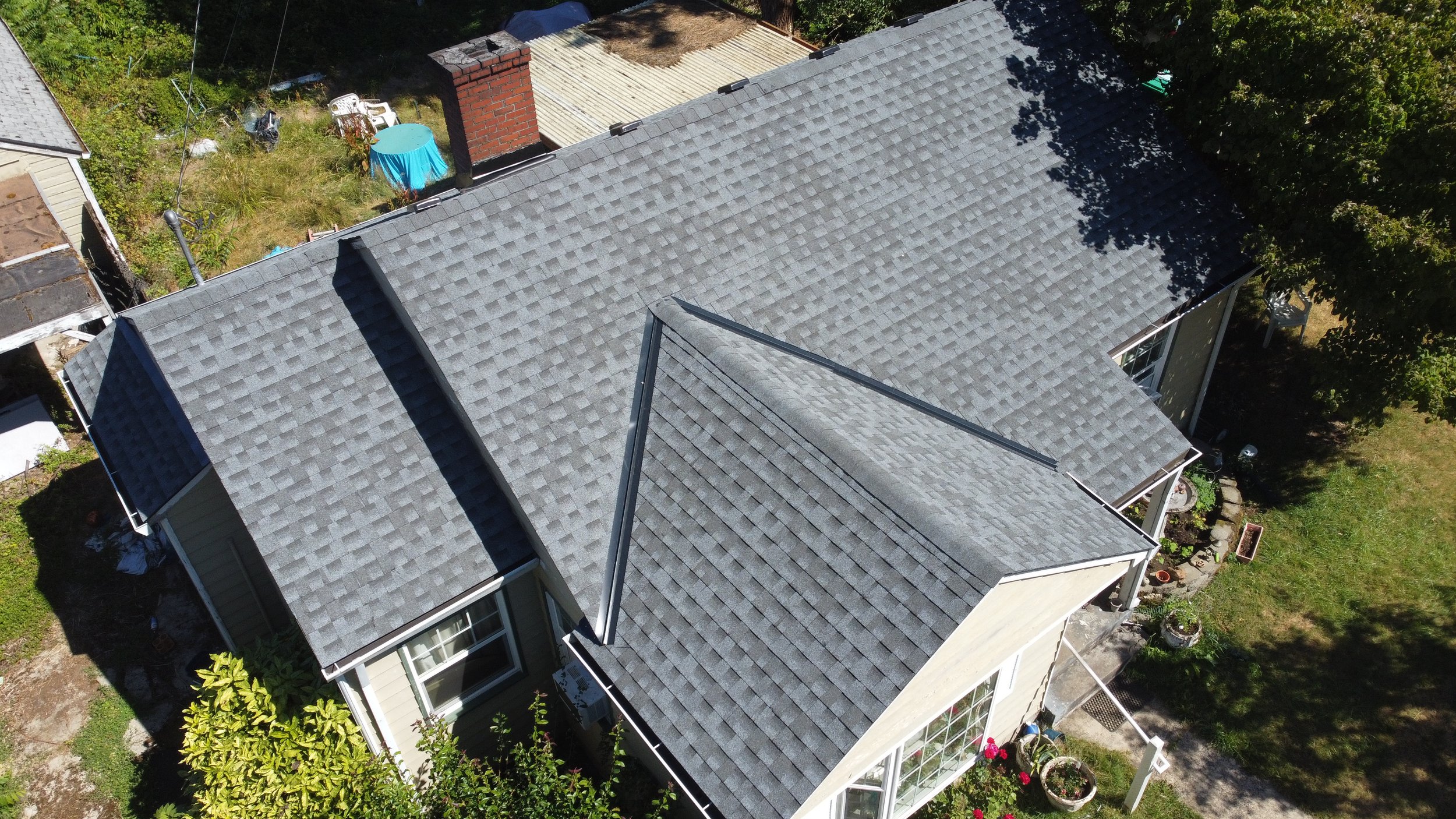Aerial view of a house with a gray shingled roof, surrounded by trees, a small backyard with a brick chimney, a patio with potted plants, and a garden with a blue table, chairs, and a garden shed.
