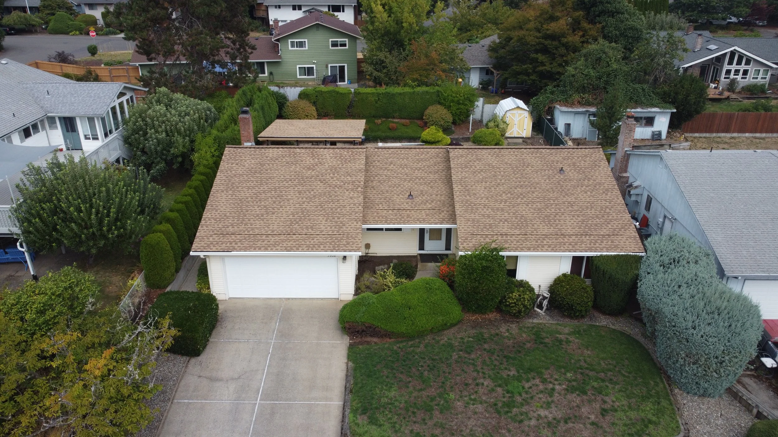 Aerial view of a suburban house with a brown shingle roof, white garage door, and front yard with mostly green grass and some bushes, surrounded by other houses and trees.