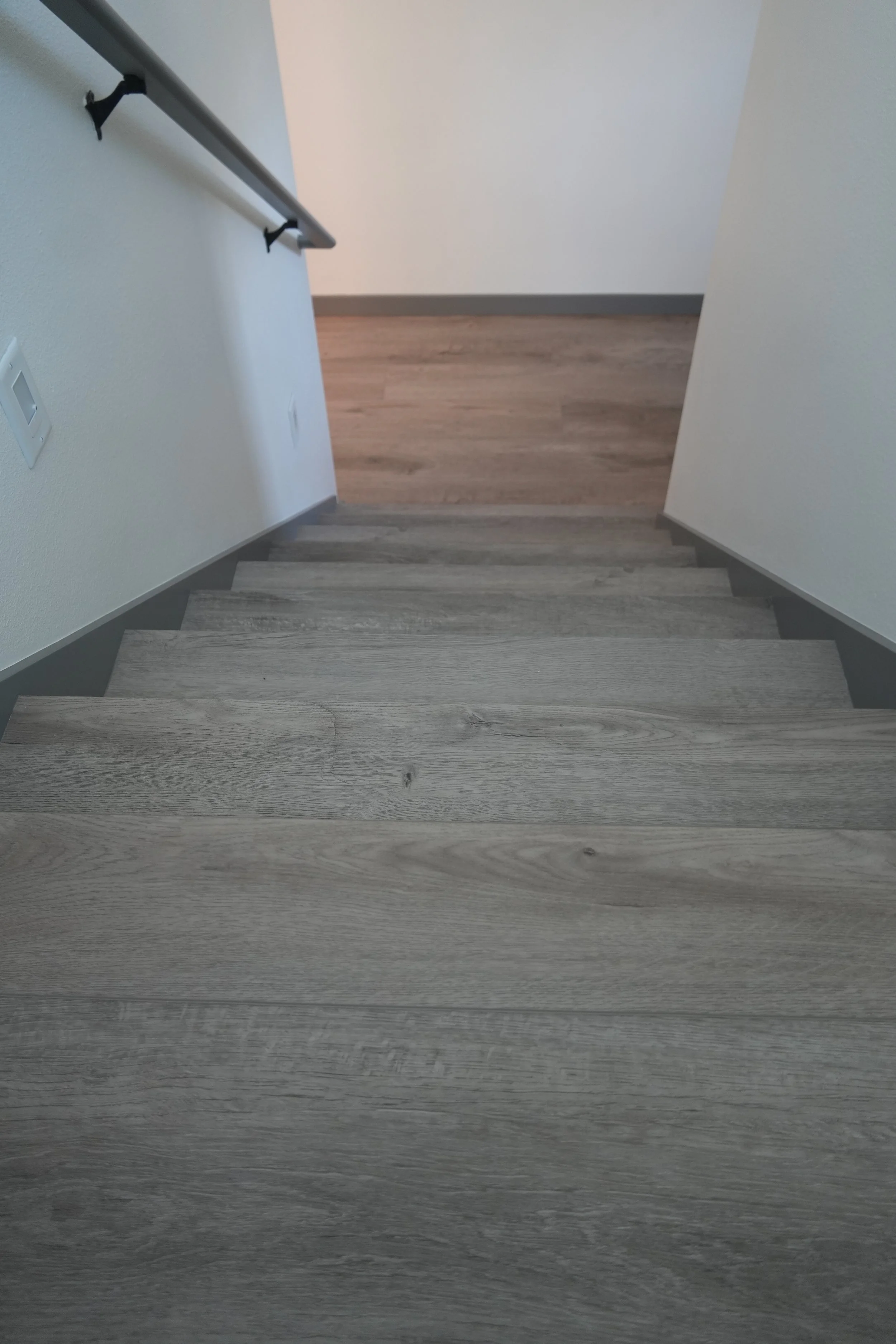 Image of a staircase with gray wooden treads, white walls, a handrail on the left, and a transition to a wood floor at the top of the stairs.