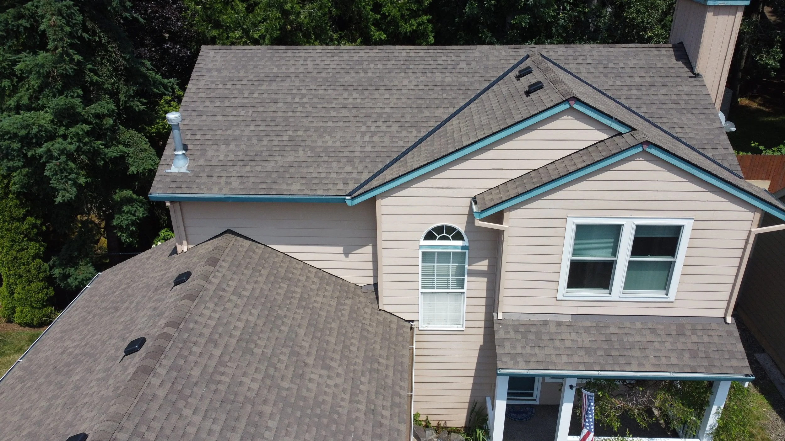 Aerial view of a house with beige siding, multiple gray shingled roof sections, and a front porch with white columns and potted plants, surrounded by green trees.