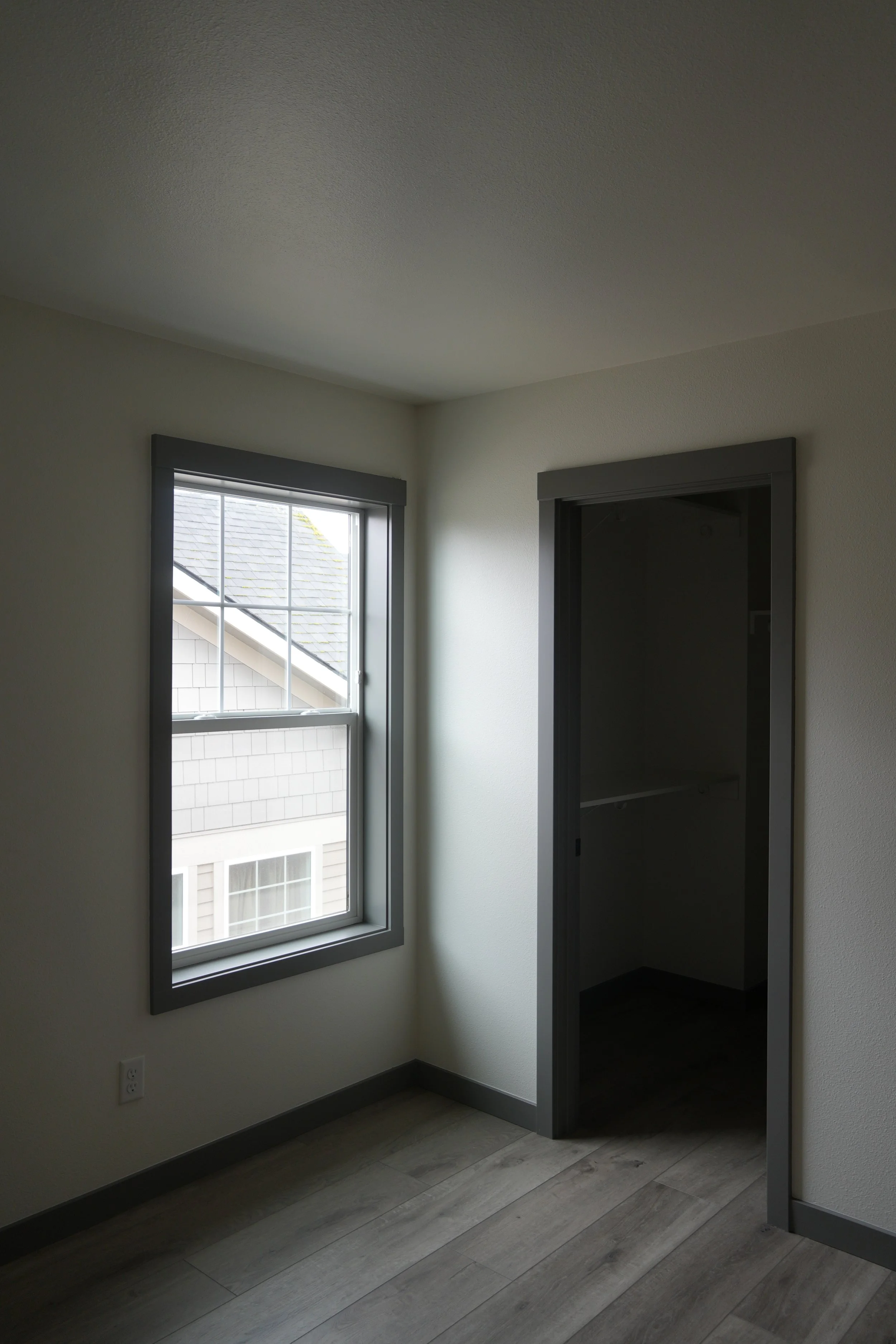 Empty room with a window and an open closet, hardwood flooring, white walls, and gray trim.