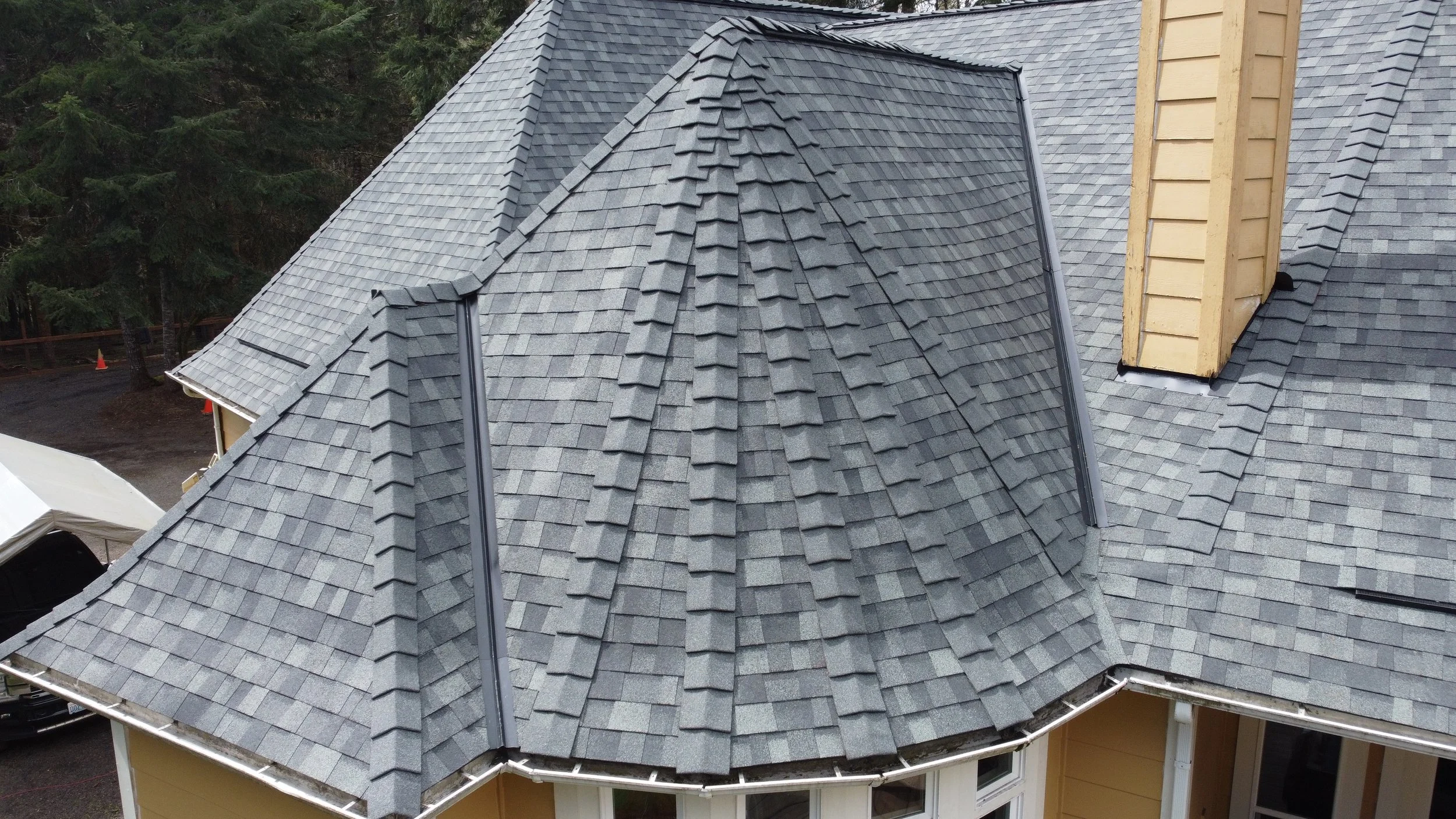 Close-up aerial view of a house roof with grey shingles, a chimney on the right, and surrounding trees in the background.