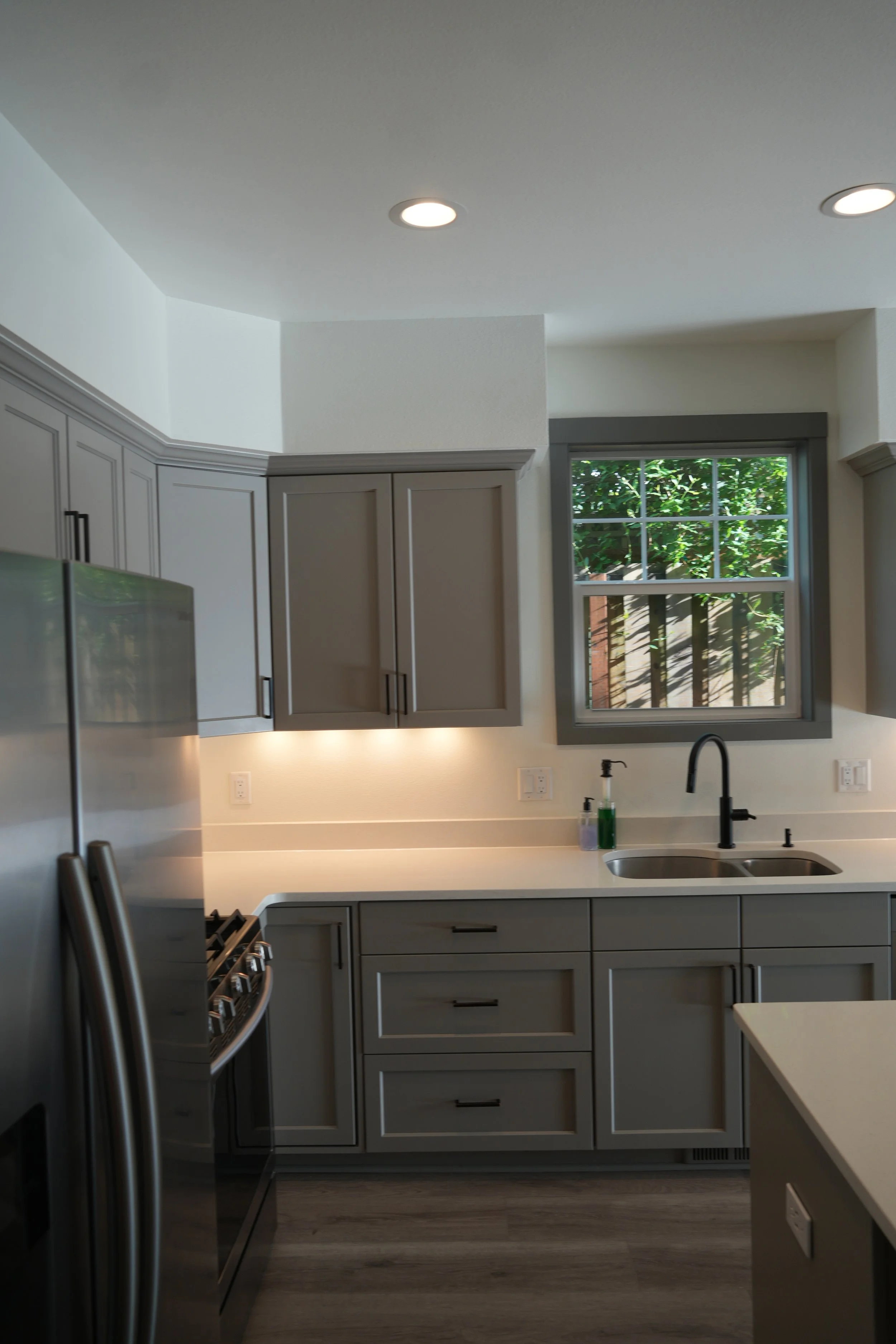 Modern kitchen with gray cabinets, stainless steel appliances, a window showing greenery outside, and under-cabinet lighting.