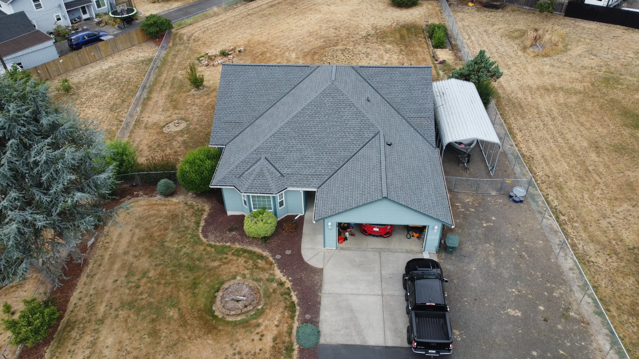 An aerial view of a suburban house with a large gray roof, a driveway with two cars, and a fenced backyard with a small covered area and some trees.