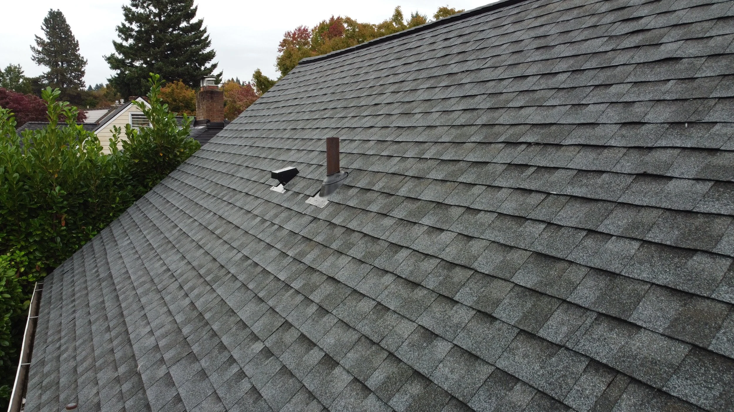 Gray shingled roof with a vent pipe and a motion-activated camera, neighboring rooftops, trees, and an overcast sky in the background.