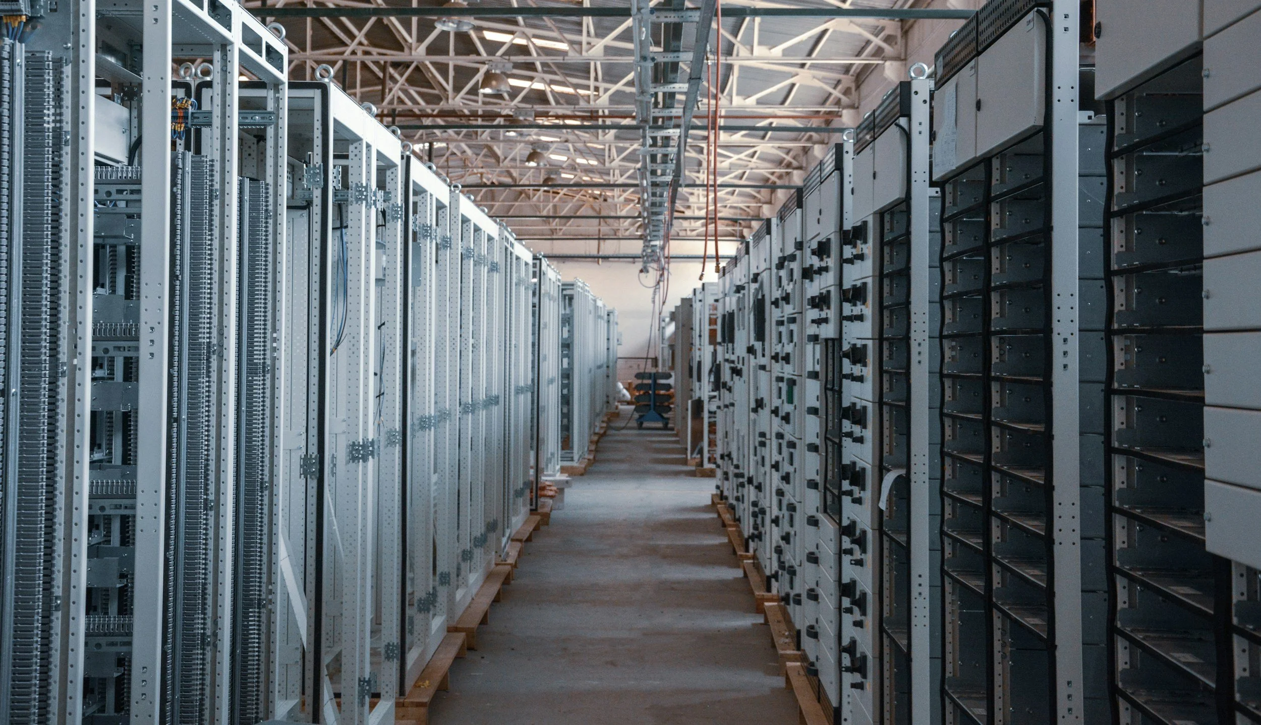 A long aisle inside a data center with server racks on both sides, some of which are empty, and exposed cables hanging from the ceiling.