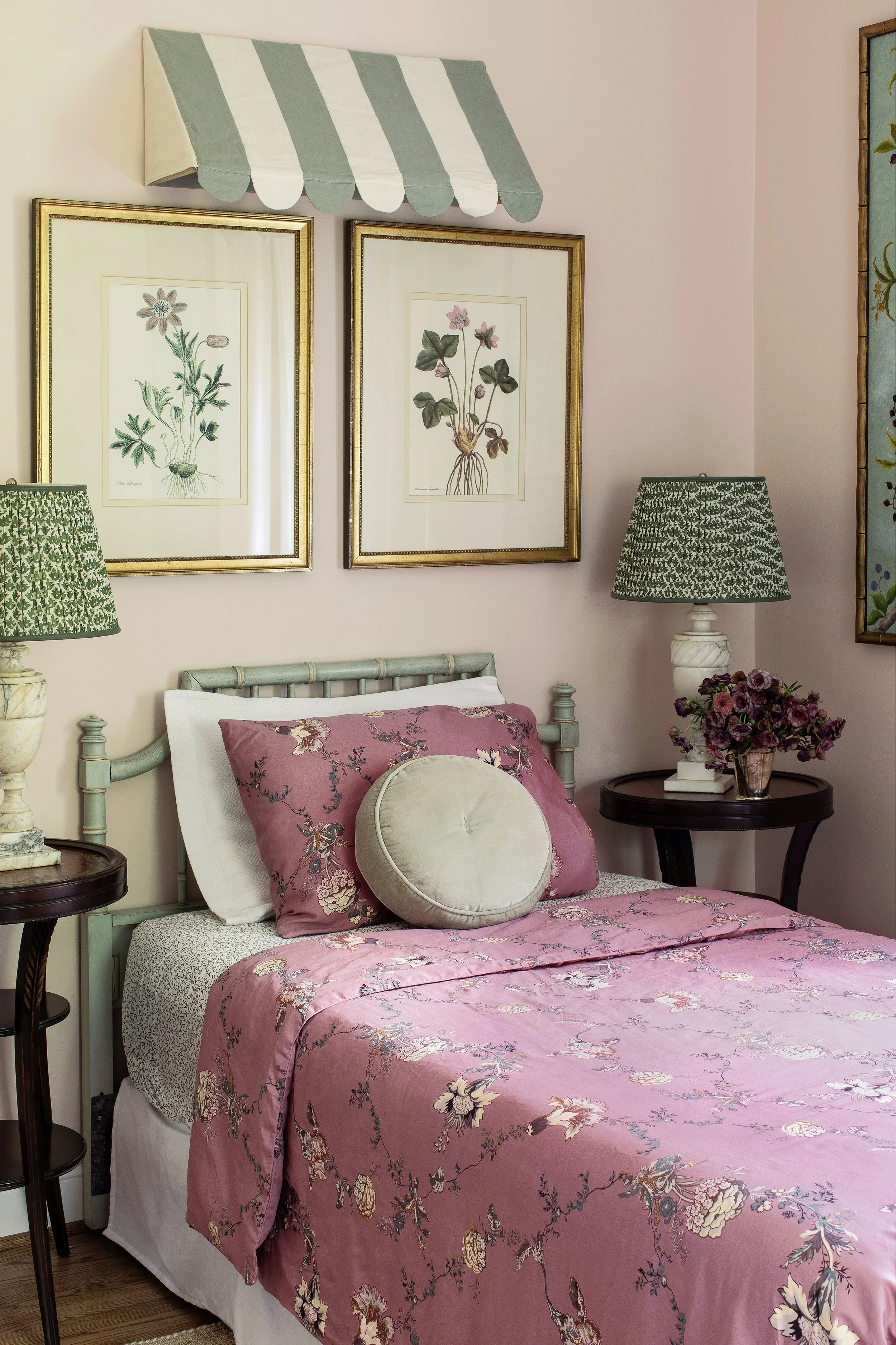 A bedroom with pink floral bedding, pale pink painted walls, two framed botanical prints on the wall, green and white striped awning above the headboard, and matching green lamps on side tables, with a bouquet of flowers on a dark wood side table.