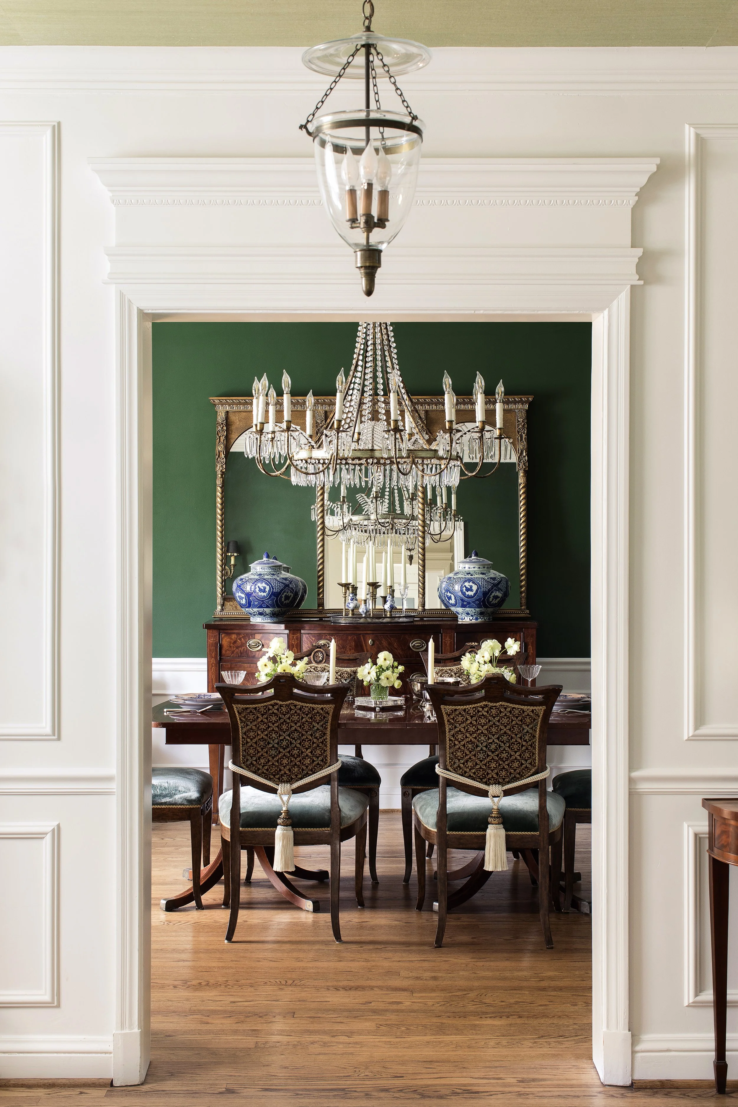 A dining room is visible through a wide doorway, featuring a dark wood table with floral centerpiece and upholstered chairs. The room has green painted walls and a large mirror above a wooden sideboard. A chandelier hangs from the ceiling.