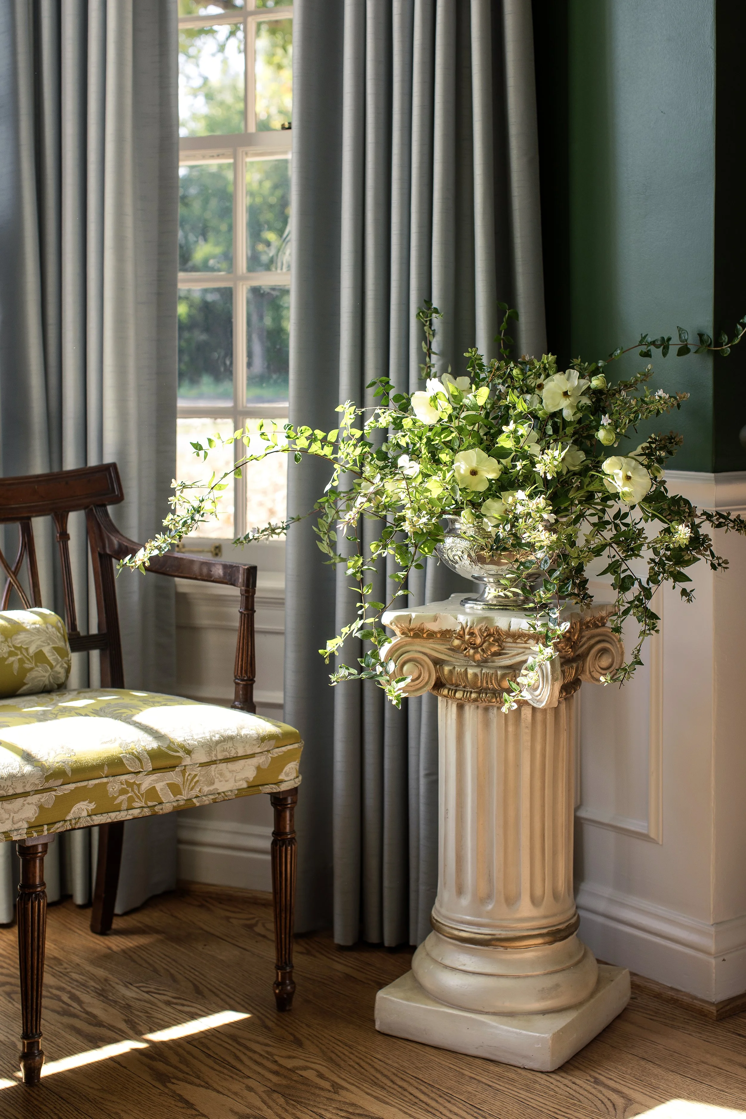 A decorative classical-style white marble pedestal with a large floral arrangement of white and green flowers in a glass vase, next to a window with grey curtains and a wood chair with a yellow patterned cushion.