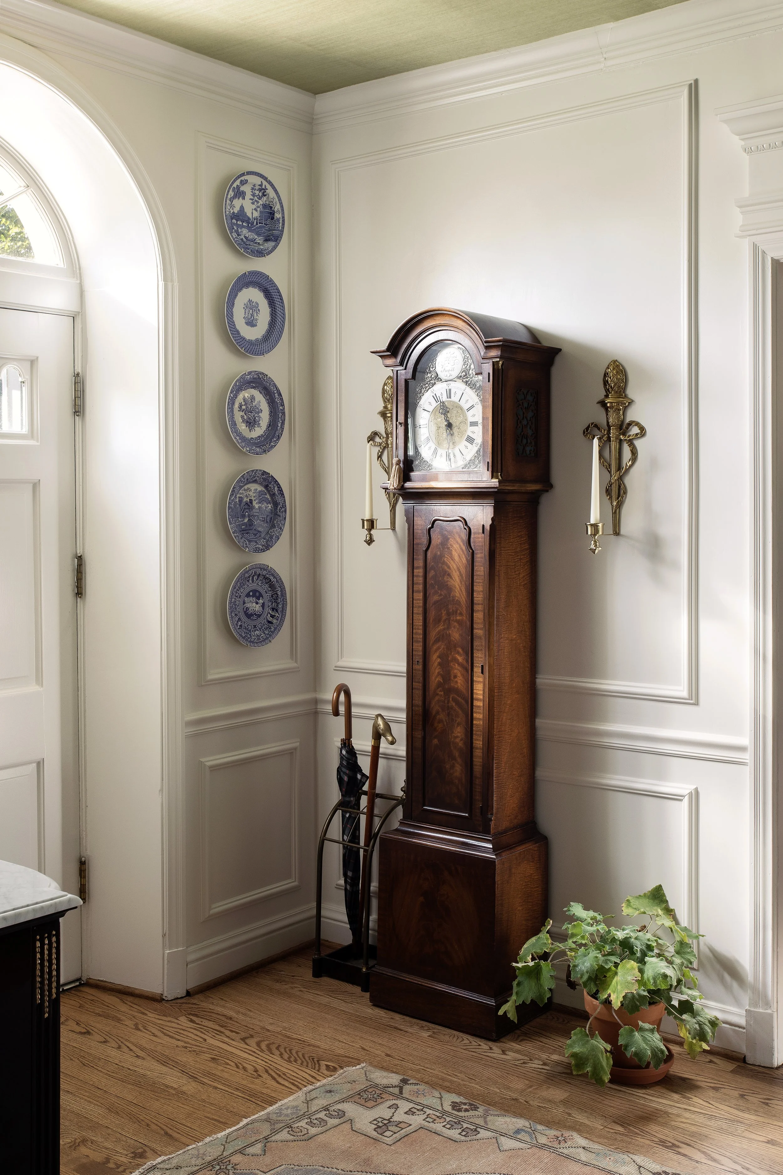 A corner of an entryway with a wooden grandfather clock, decorative blue and white wall plates, a coat rack with umbrellas, and a potted green plant on a hardwood floor with a patterned rug. Light from a nearby window illuminates the space.