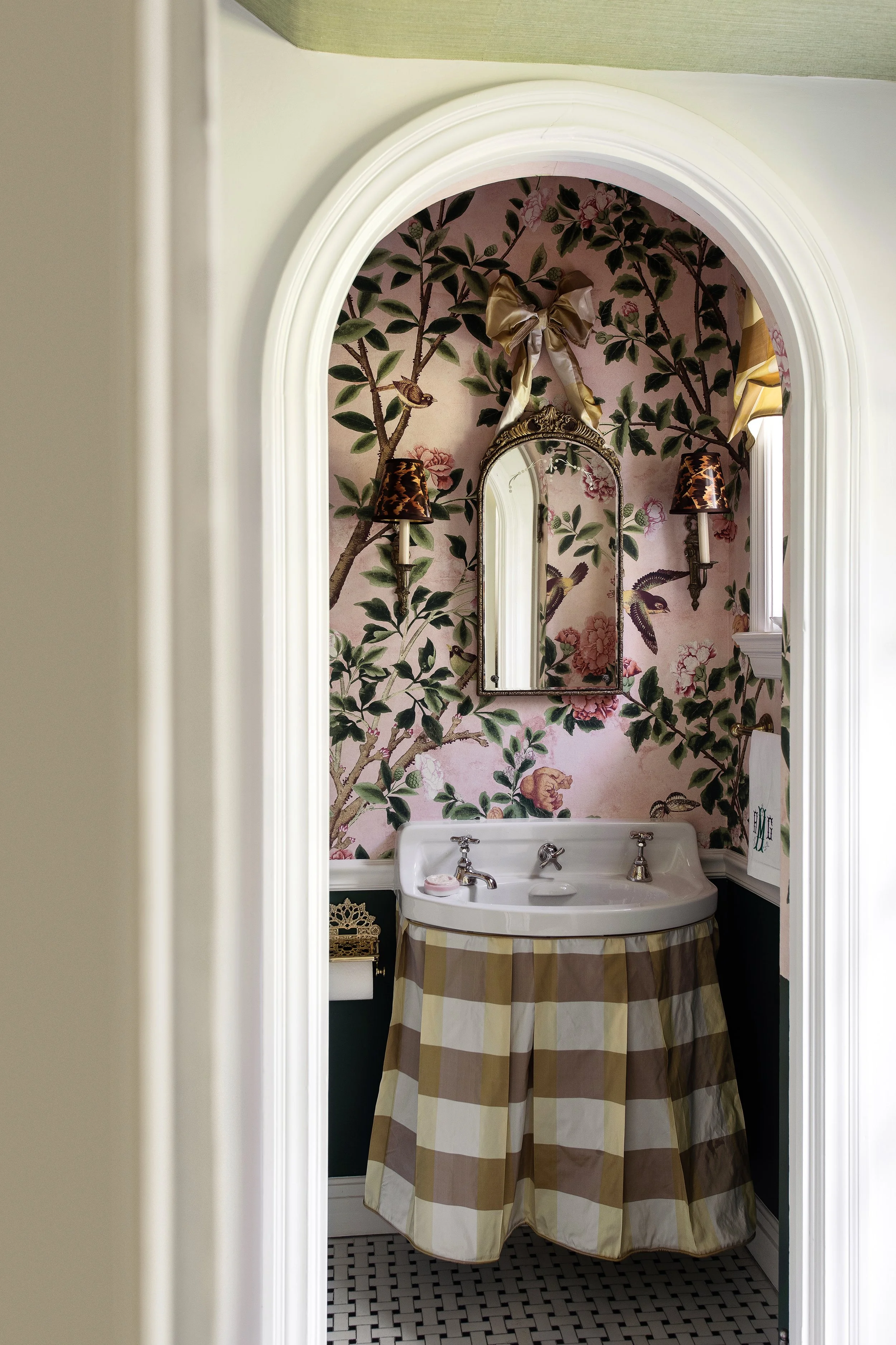 A powder room with floral wallpaper, a vintage sink with a striped fabric skirt, a mirror with gold trim, and twin wall sconces with animal-print shades.