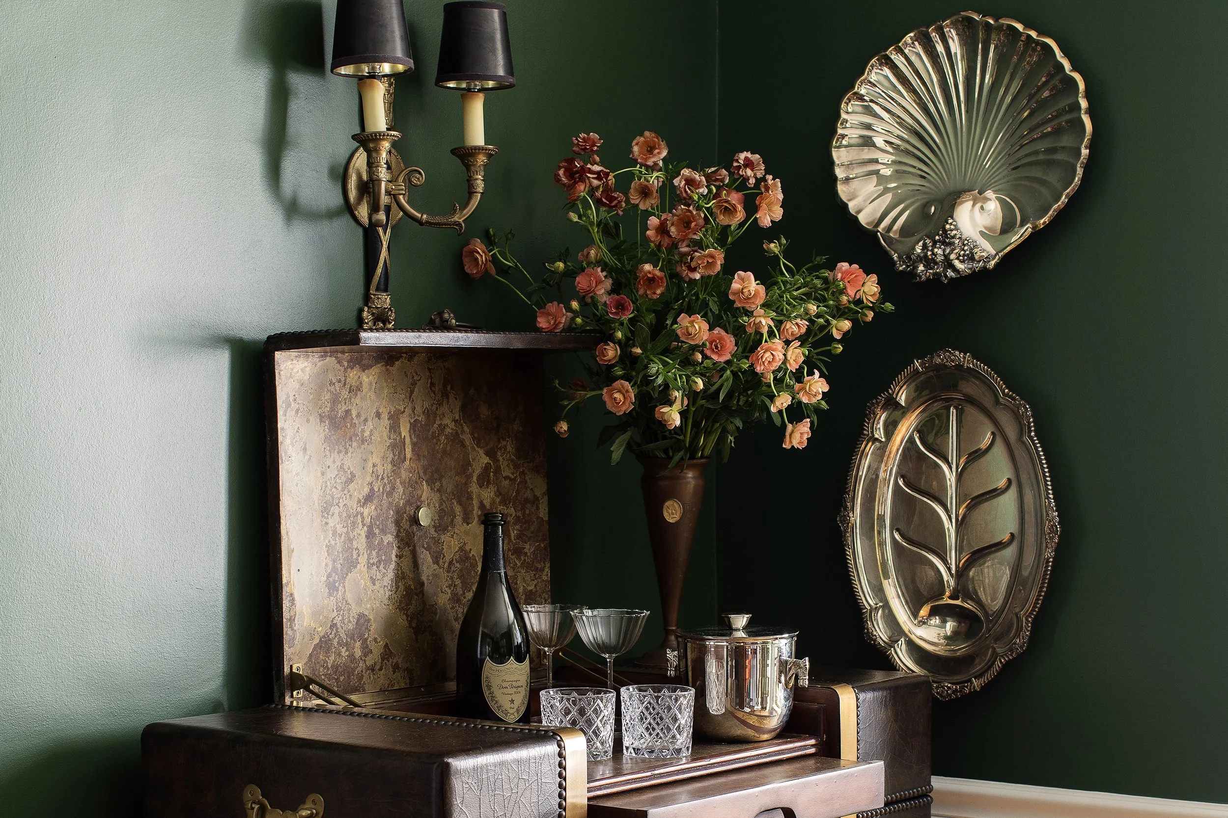 A vintage dark wood bar furnishing is decorated with glassware and a bouquet of flowers in a vase. Walls are painted green. Silver shell-shaped molds and a large silver tray with a leaf design are seen on the wall.
