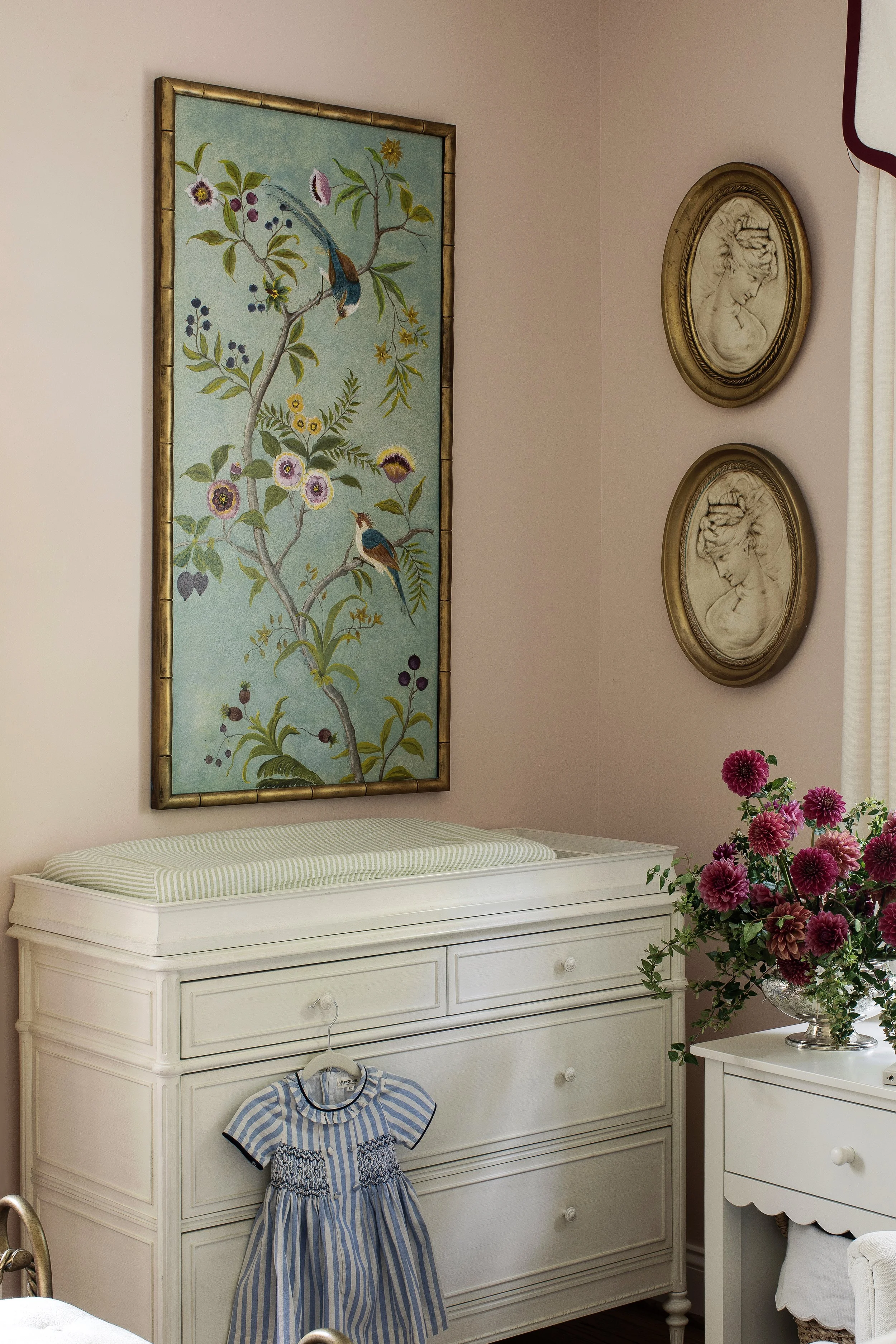A white dresser with drawers and a children's dress hanging on the handle. Next to it is a small white nightstand with a silver vase holding flowers. On the wall above, there is a framed tapestry with a bird and flowers design, and two classical style portraits in gold frames. The room has light pink walls and a window with a cream curtain edged in dark red.