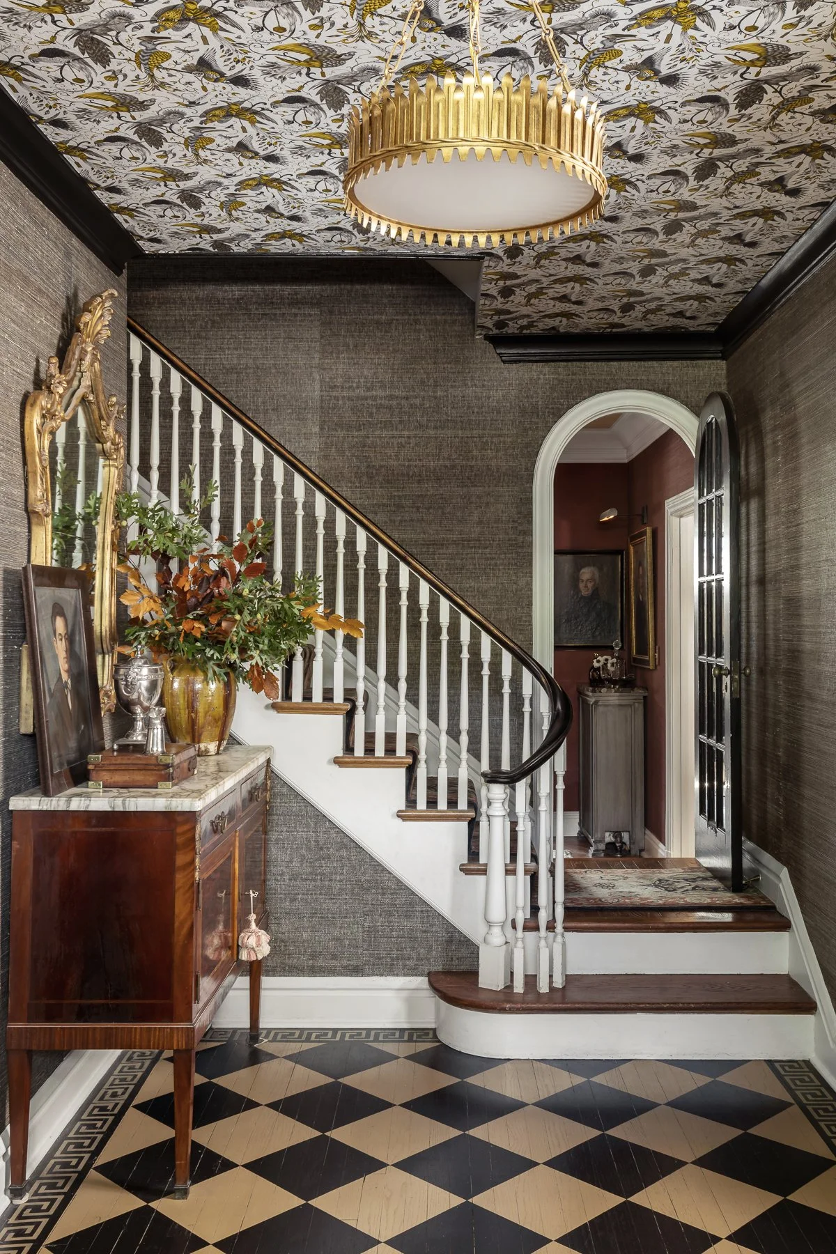 A foyer with a staircase featuring a dark wood handrail, a black and beige checkered floor, dark textured wallpaper, a gold chandelier, a wooden sideboard with a marble top, a painting, a large oval mirror, a vase, and framed artwork on the walls.