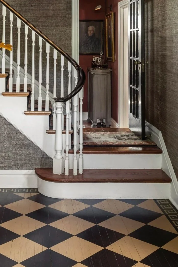 Interior view of a home entryway featuring a staircase with white spindles and dark wood handrail, a black and cream tiled floor, and a small hallway with artwork and a gray cabinet.