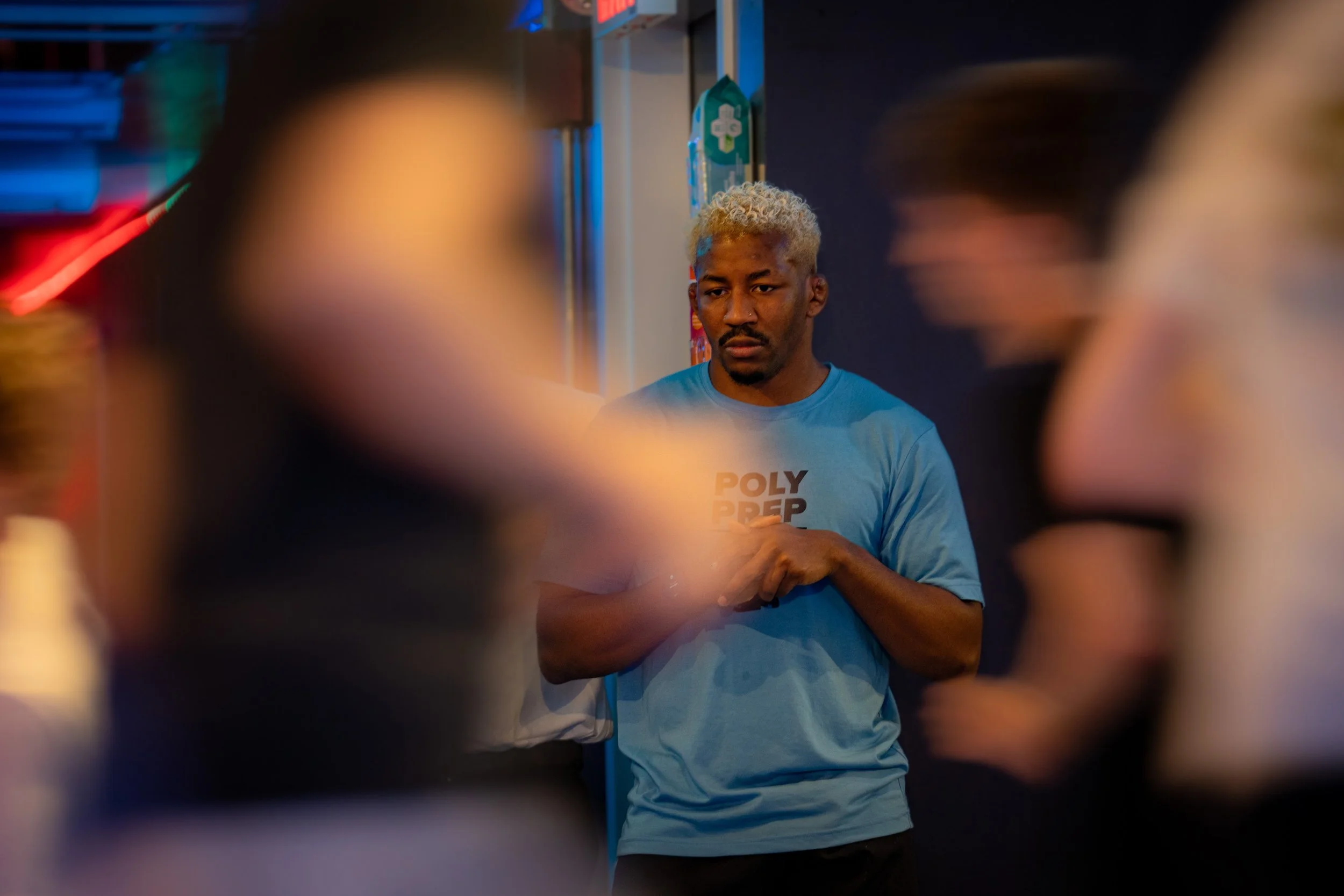 A young man with bleached blond hair and a blue shirt stands in a gym or fitness studio, focused and serious, surrounded by blurred people in motion.
