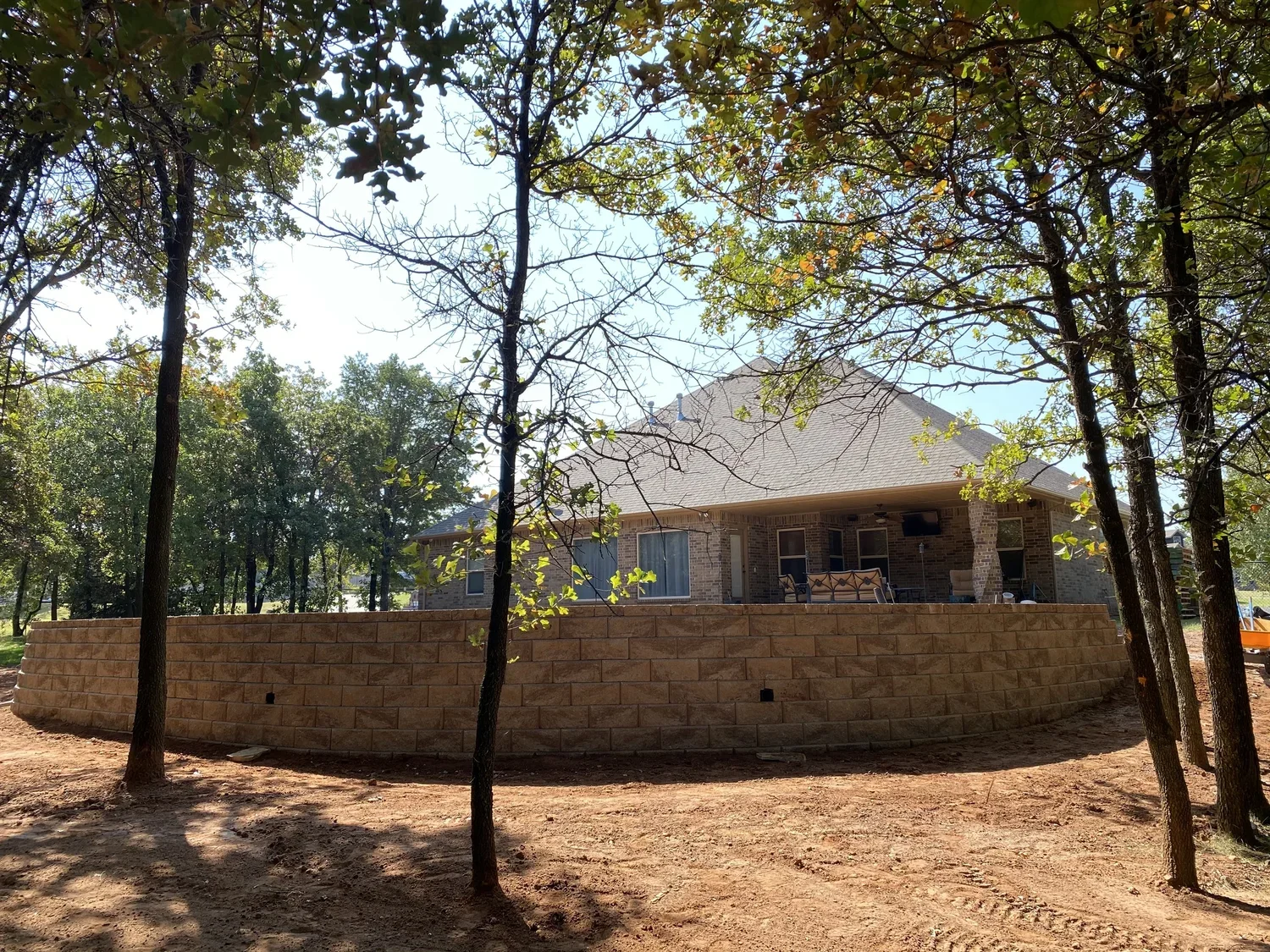 A house with a brick exterior, a sloped roof, and a raised patio enclosed by a brick wall. There are trees with leaves partially shading the house and a dirt ground in the foreground.