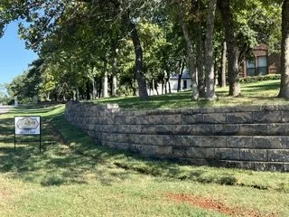 A landscaped area with a curved stone wall, green grass, and tall trees in the background.