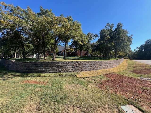 A small raised landscaped area with grass and trees, surrounded by a brick wall, located beside a paved street under a clear blue sky.