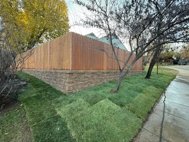 A wooden backyard fence with stone foundation, trees with sparse leaves, green grass, and a sidewalk in a suburban neighborhood.