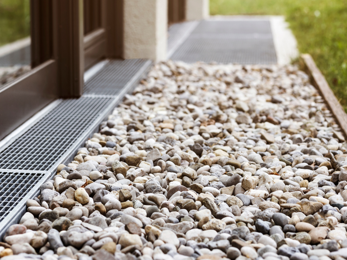 Close-up view of a gravel pathway with a metal grate sidewalk next to a building, surrounded by grass.