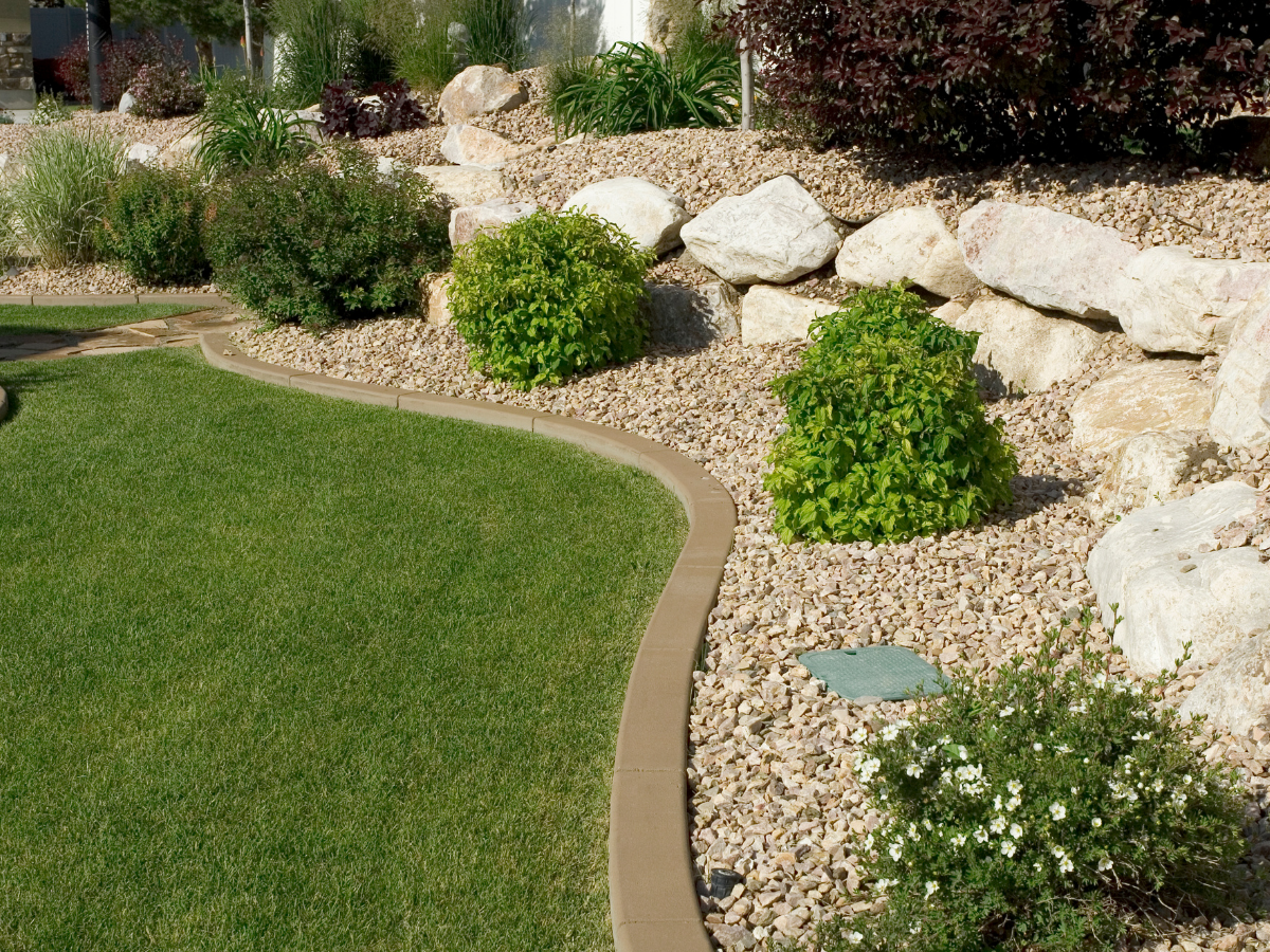 Landscaped backyard with green grass, a curved stone border, and a rock garden with shrubs and plants, including small bushes, large rocks, and decorative gravel.