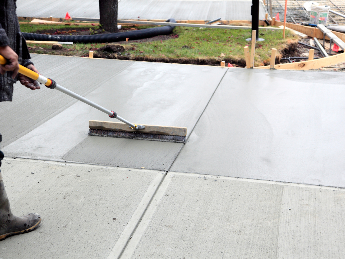 Person using a push broom to smooth fresh concrete sidewalk at a construction site.