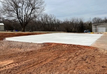 Empty parking lot with a concrete slab, surrounded by dirt and sparse trees in the background.