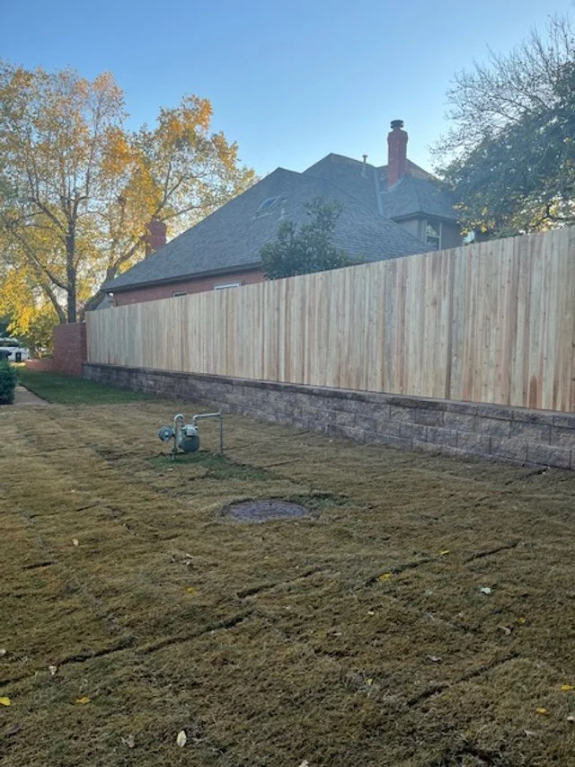 View of a backyard with freshly laid sod, a utility meter, a brick wall, and a tall wooden fence. In the background, there is a house with a steep roof and a chimney, along with trees with yellow and orange leaves against a clear blue sky.