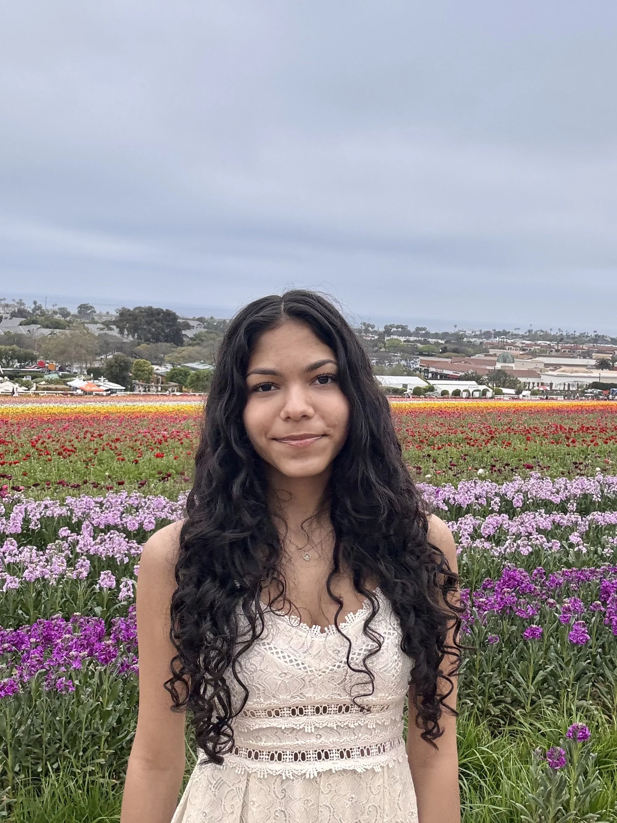 A young woman with long curly black hair wearing a cream-colored lace dress standing in front of flower fields with pink, purple, red, and orange flowers under a cloudy sky. She's had success with Reframe CBT