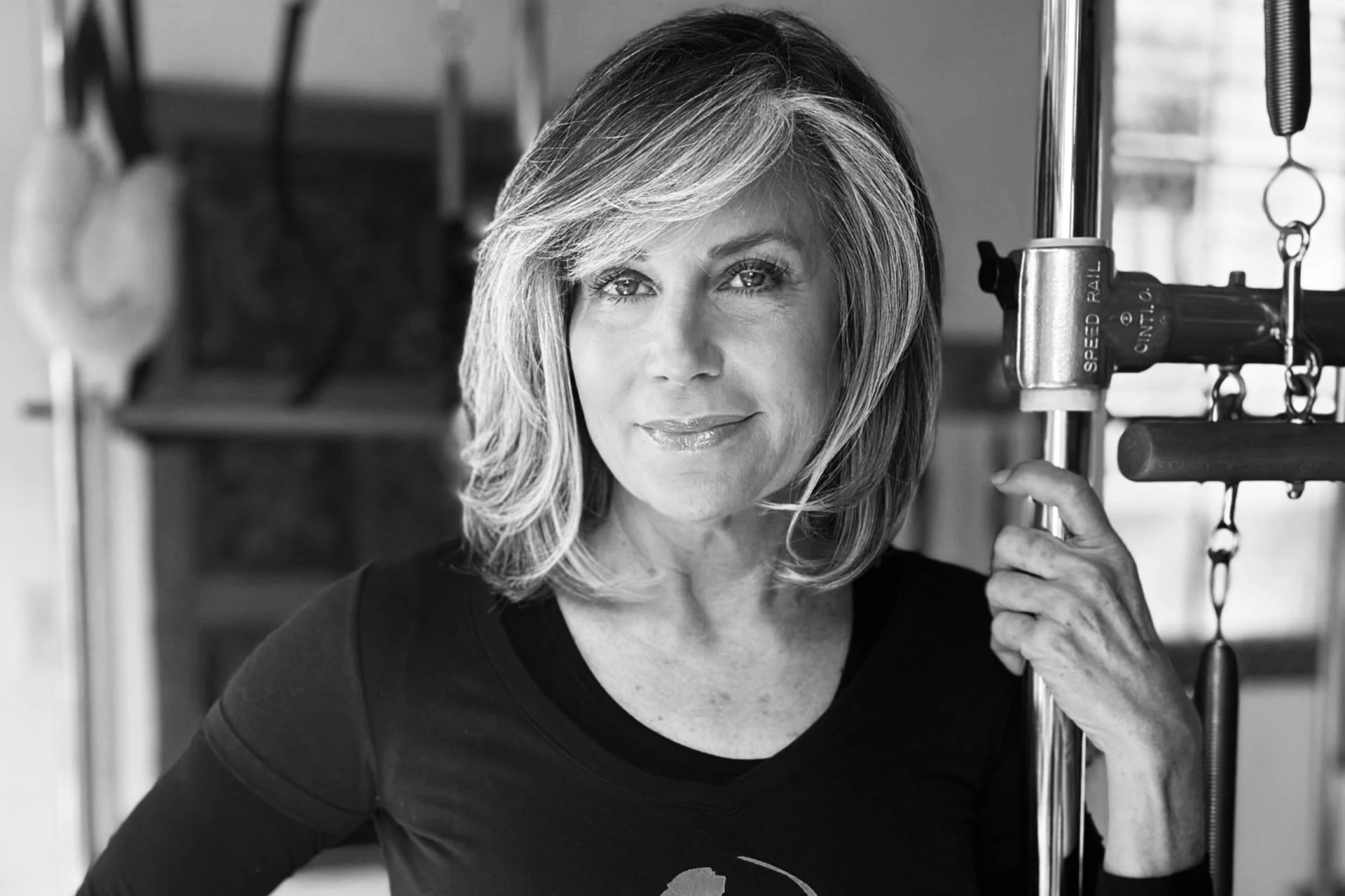 A black and white portrait of a middle-aged woman with shoulder-length hair, standing in a gym, holding onto gym equipment, with a confident expression.