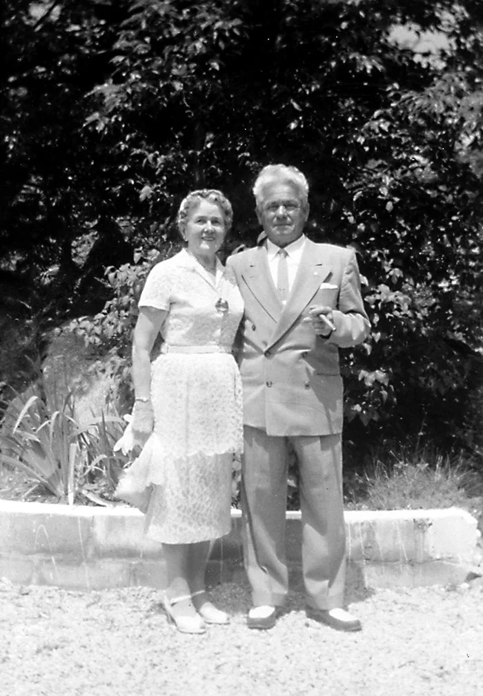 A black and white photo of a woman in a dress and a man in a suit standing outdoors in front of trees.