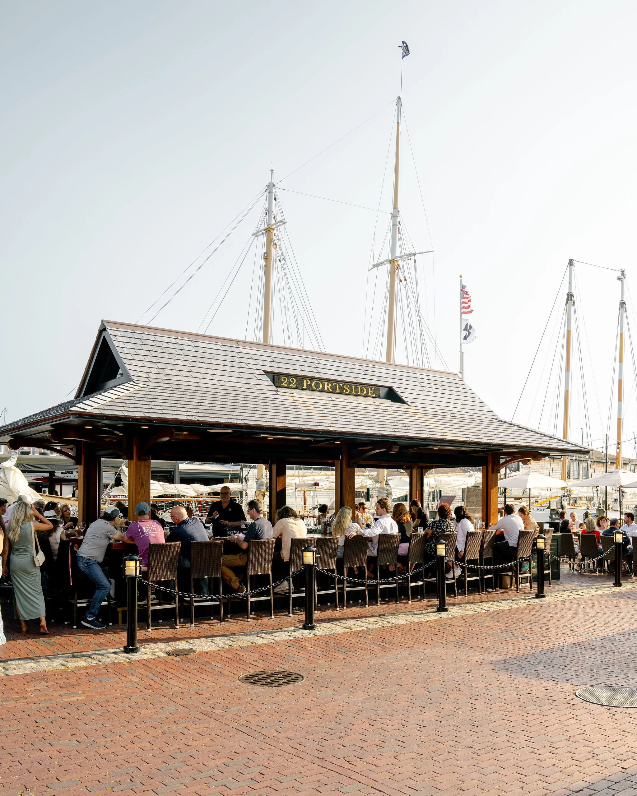 People dining on outdoor patio at Portside restaurant with sailboats docked in the background.